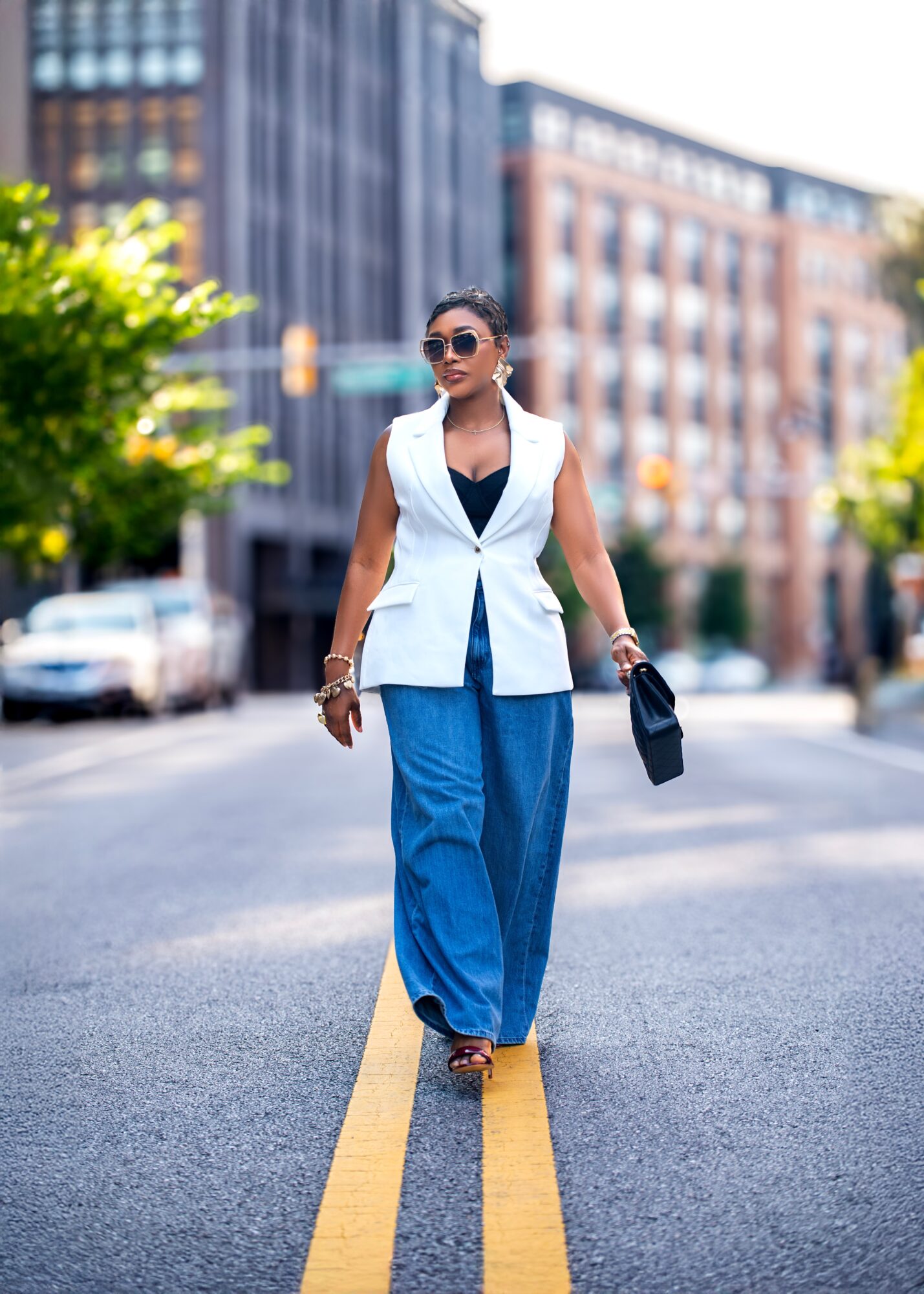 Woman walking on city street wearing sunglasses, sleeveless white blazer, black top, and blue wide-leg pants, holding a black bag.