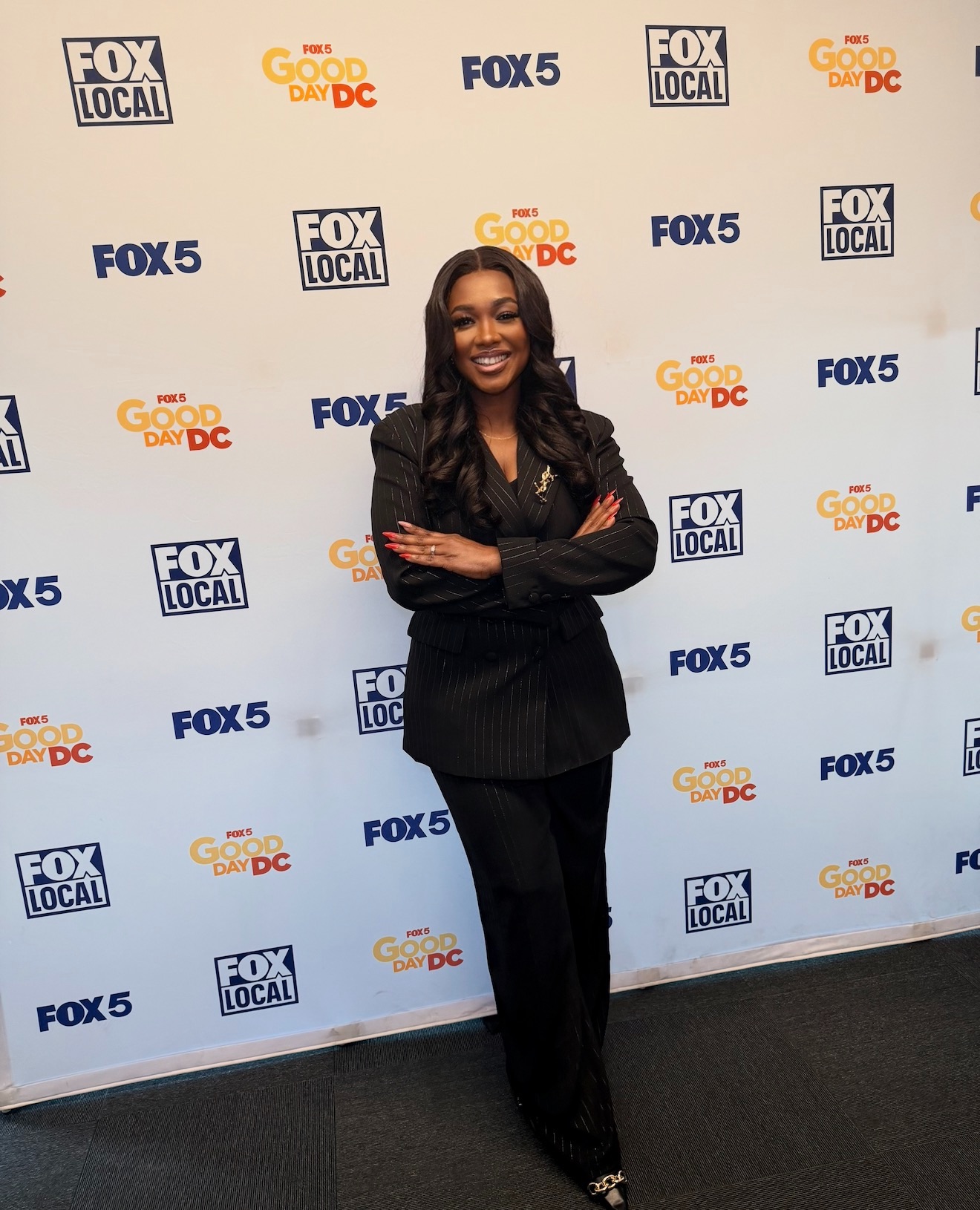 Woman in black suit standing with arms crossed, smiling in front of a backdrop with TV network logos.
