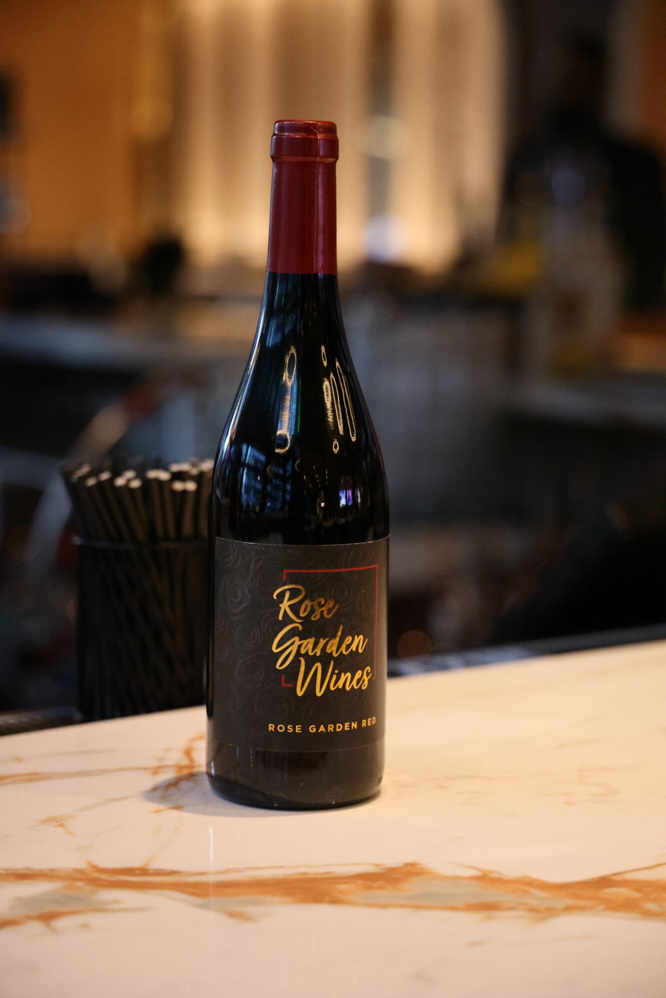 A bottle of red wine on a table with a blurred background, black label, and red cap.