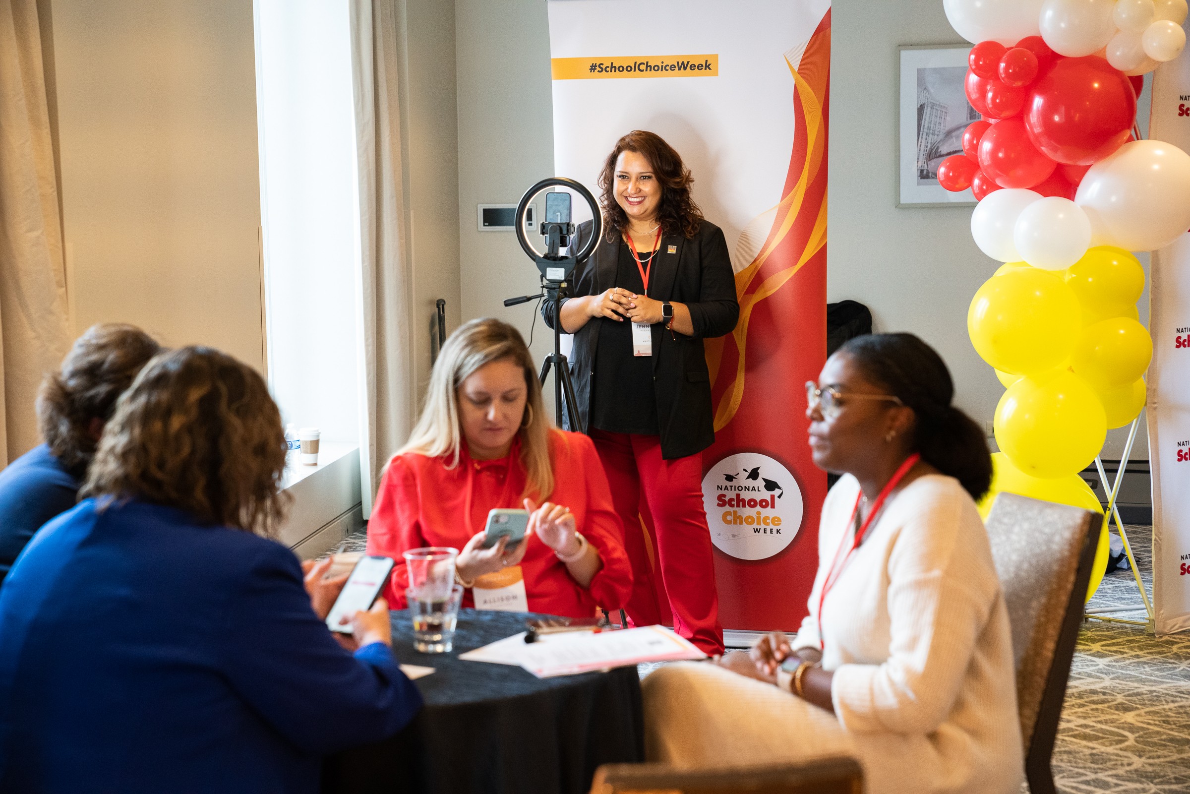 Group of women sitting at a table in a conference room, with a woman standing near a banner and balloons, holding a microphone.
