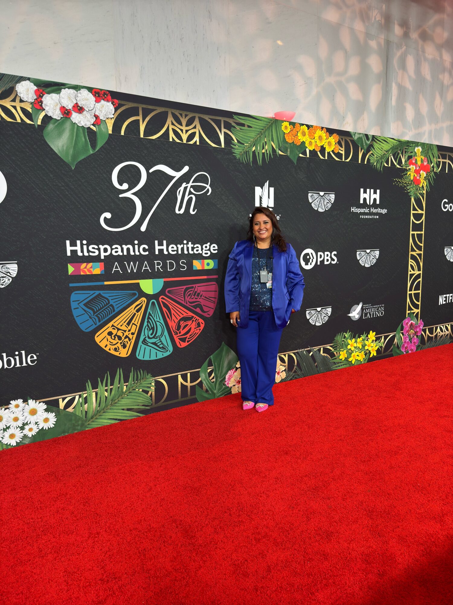 Woman in blue suit standing on red carpet in front of a black backdrop with event logos and floral decorations.