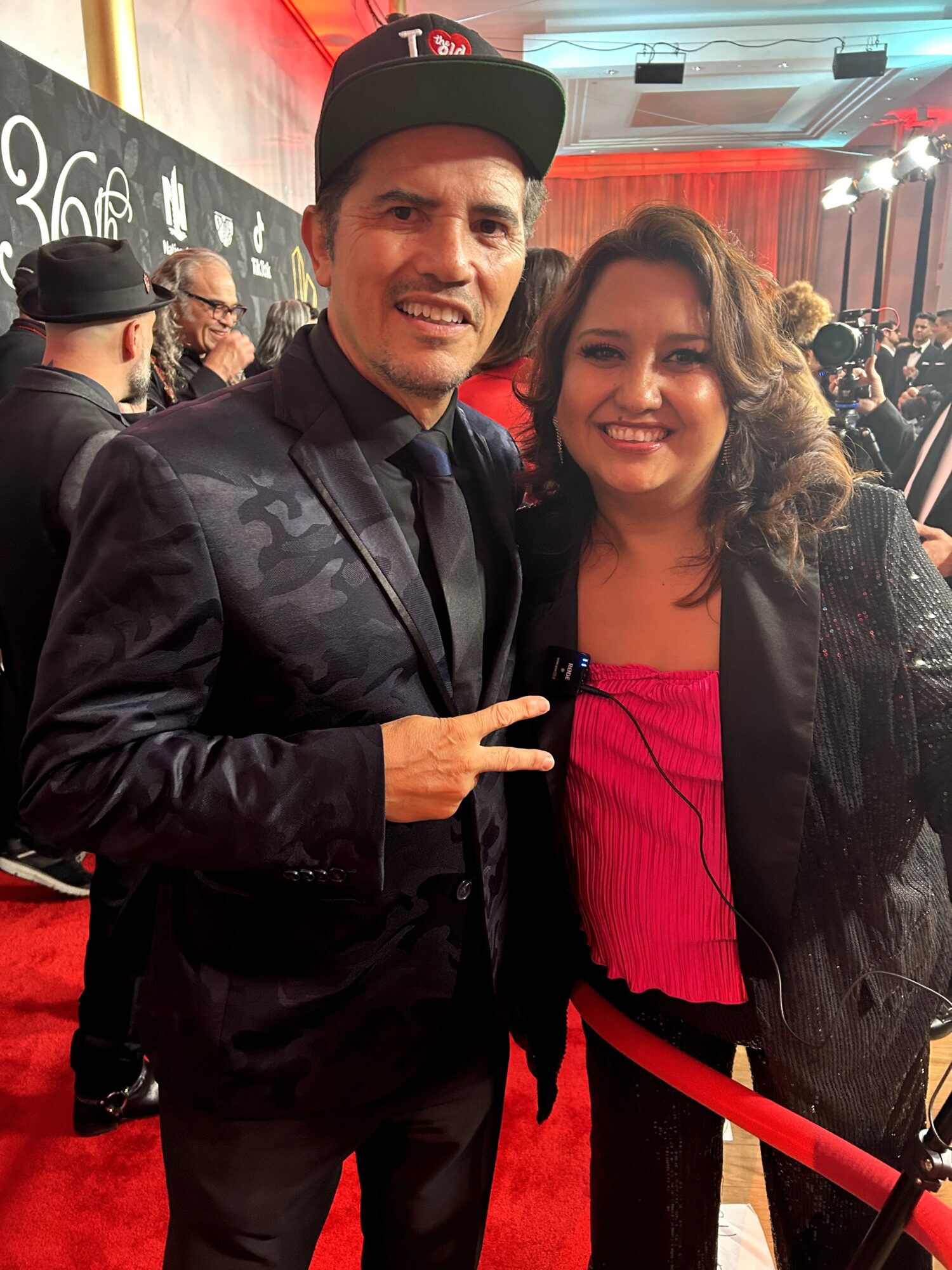 Man and woman smiling at an event, the man making a peace sign, crowd in background, red carpet, indoor setting.