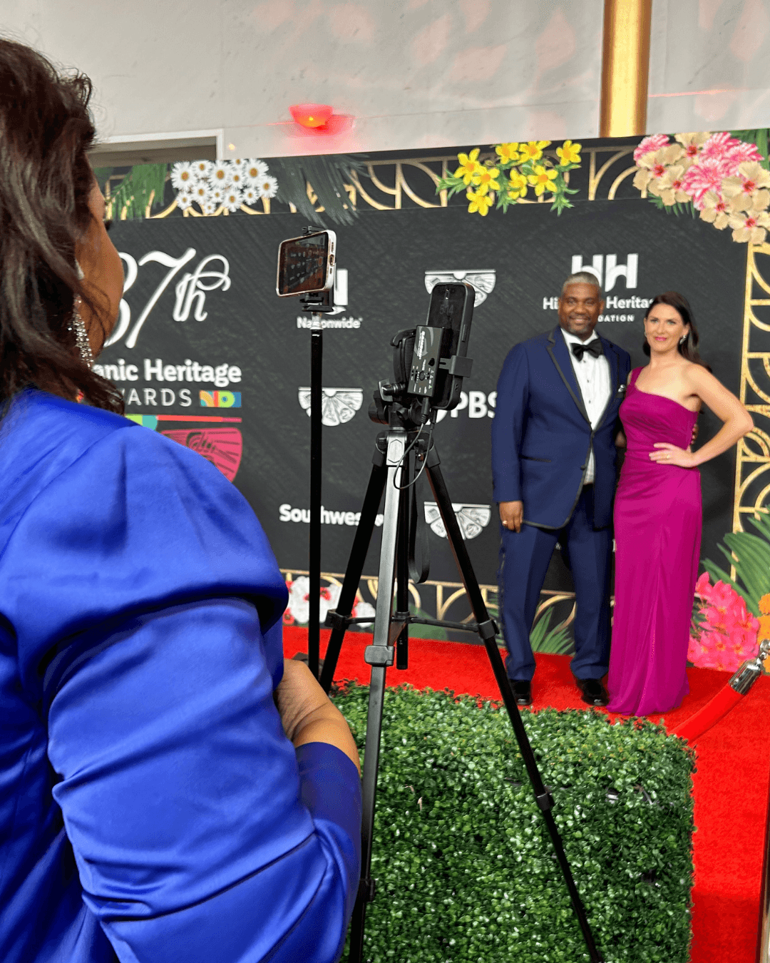 Person in blue dress taking a photo of a man in a tuxedo and woman in a pink gown on a red carpet, backdrop with event logos.