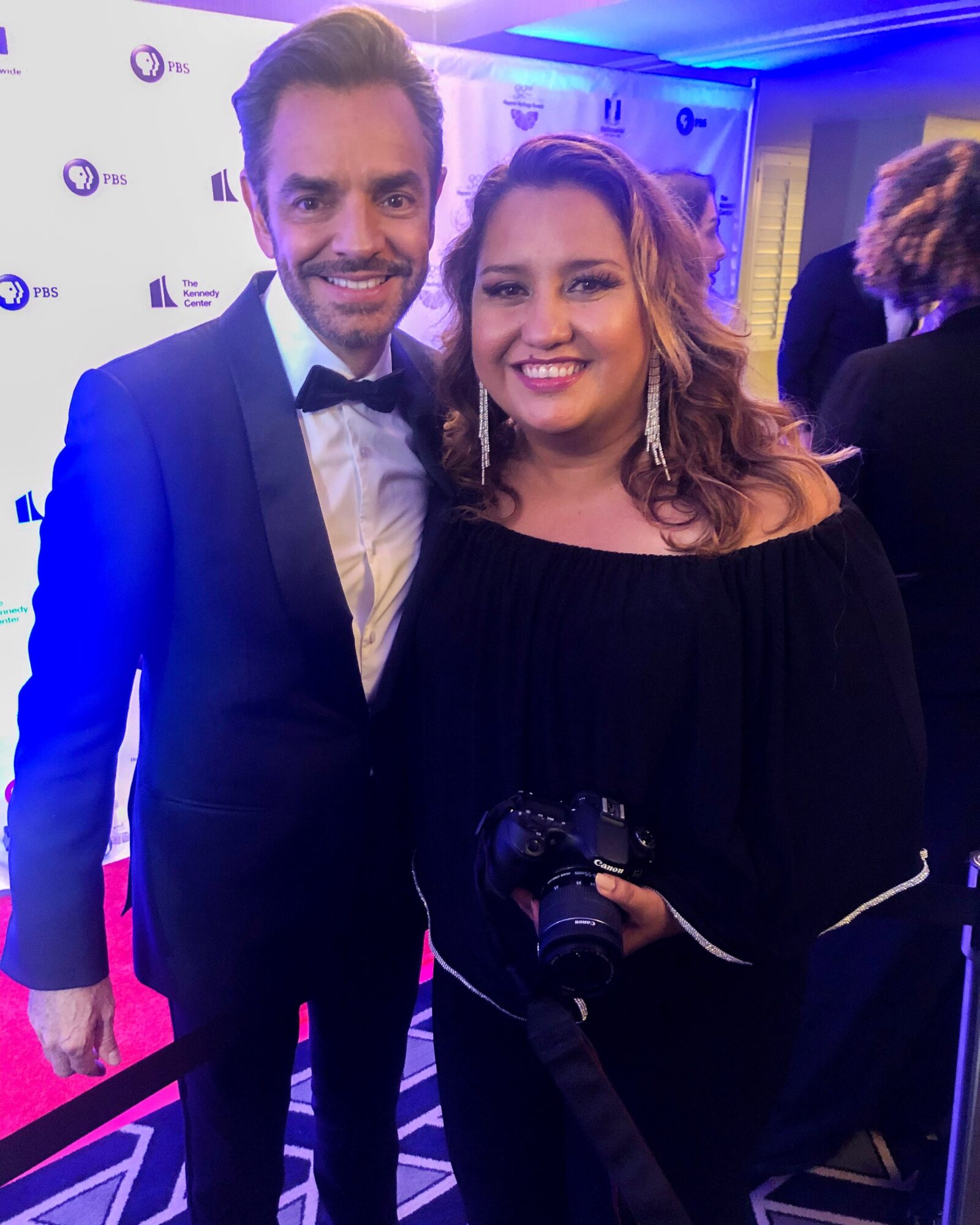 Man in tuxedo and woman in black dress smiling at event, holding a camera, with a patterned floor and blue lighting.
