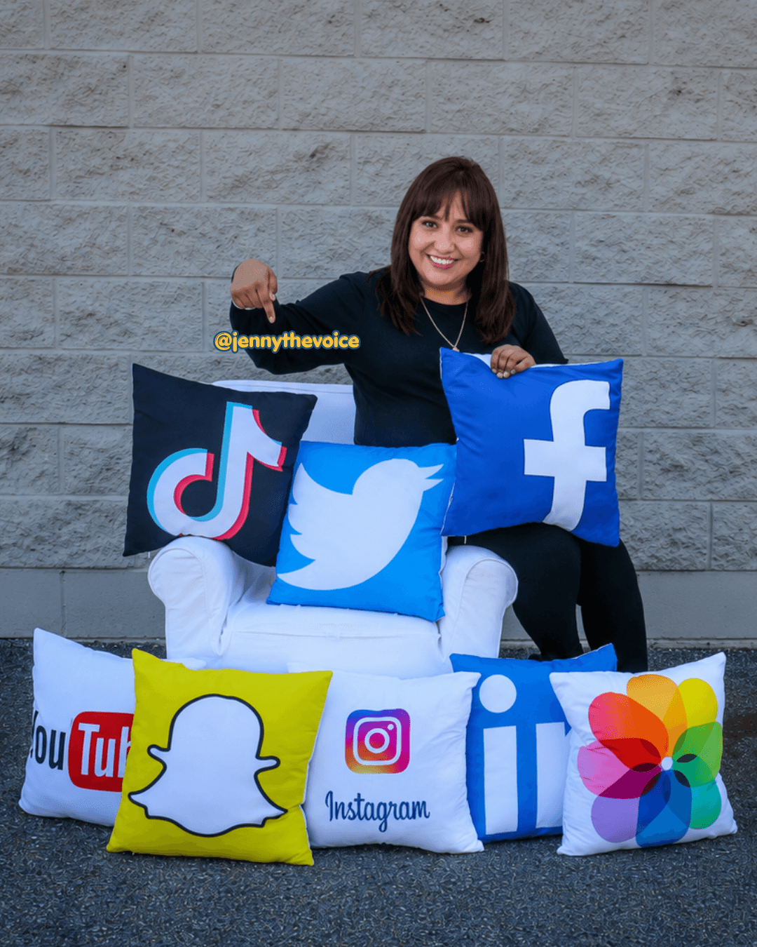 Woman sitting behind social media pillows, smiling, against a gray brick wall, with TikTok, Twitter, Facebook, Snapchat, Instagram, YouTube, LinkedIn, and rainbow-colored pillows.