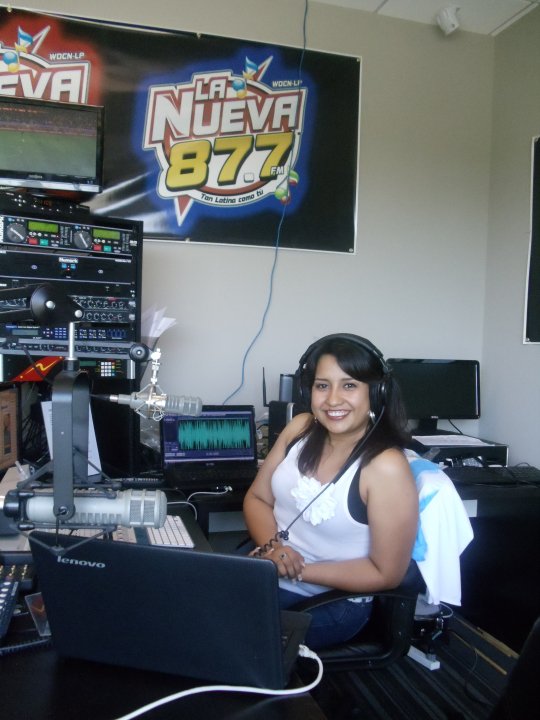 Smiling woman with headphones sitting at a radio station desk with broadcasting equipment and a sign reading 'La Nueva 877'.