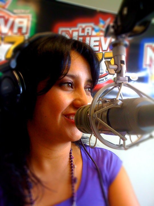 Woman with dark hair speaking into a microphone in a radio studio, wearing headphones and a purple top.