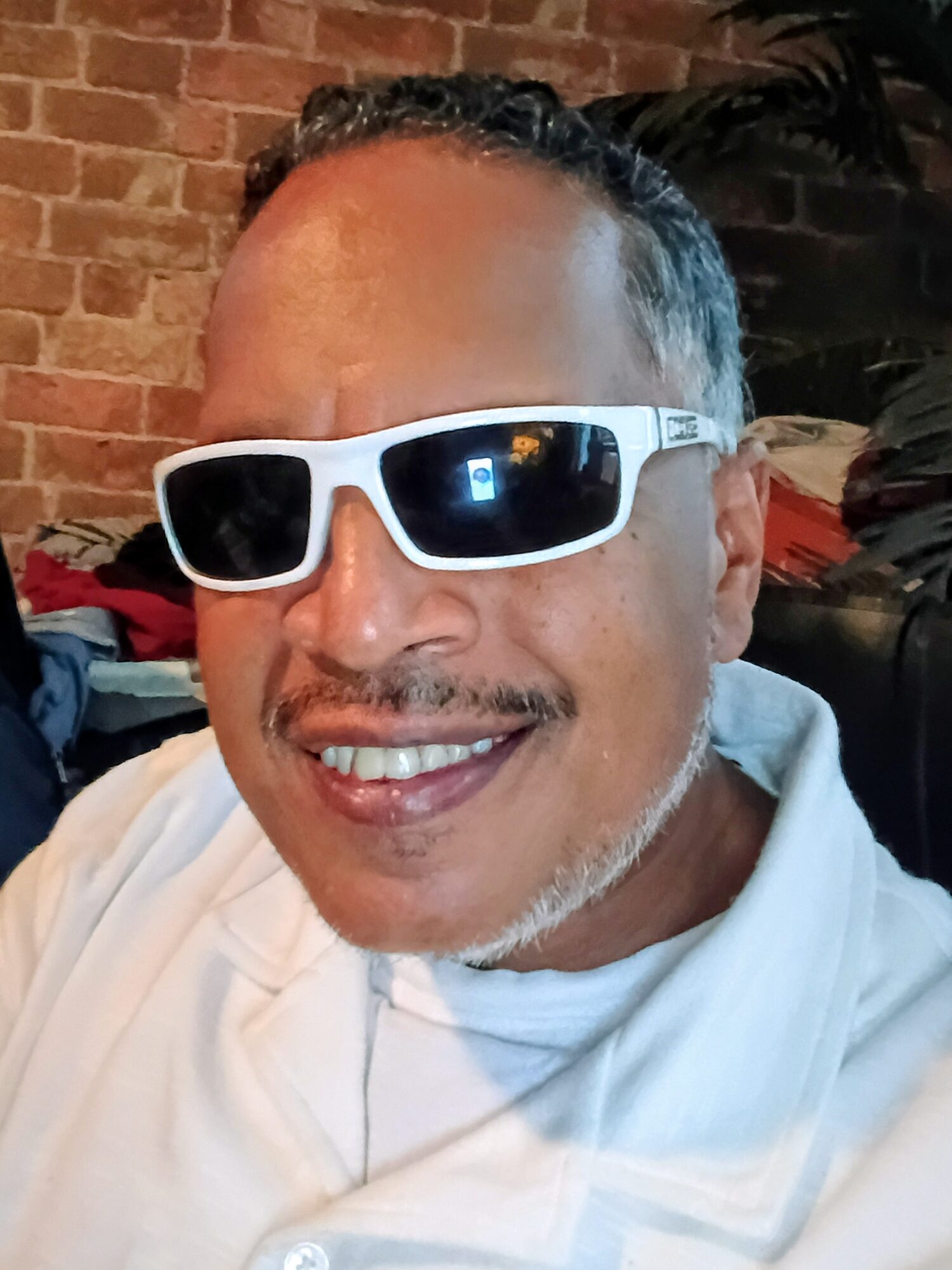Man smiling wearing white sunglasses and a white shirt, brick wall background.