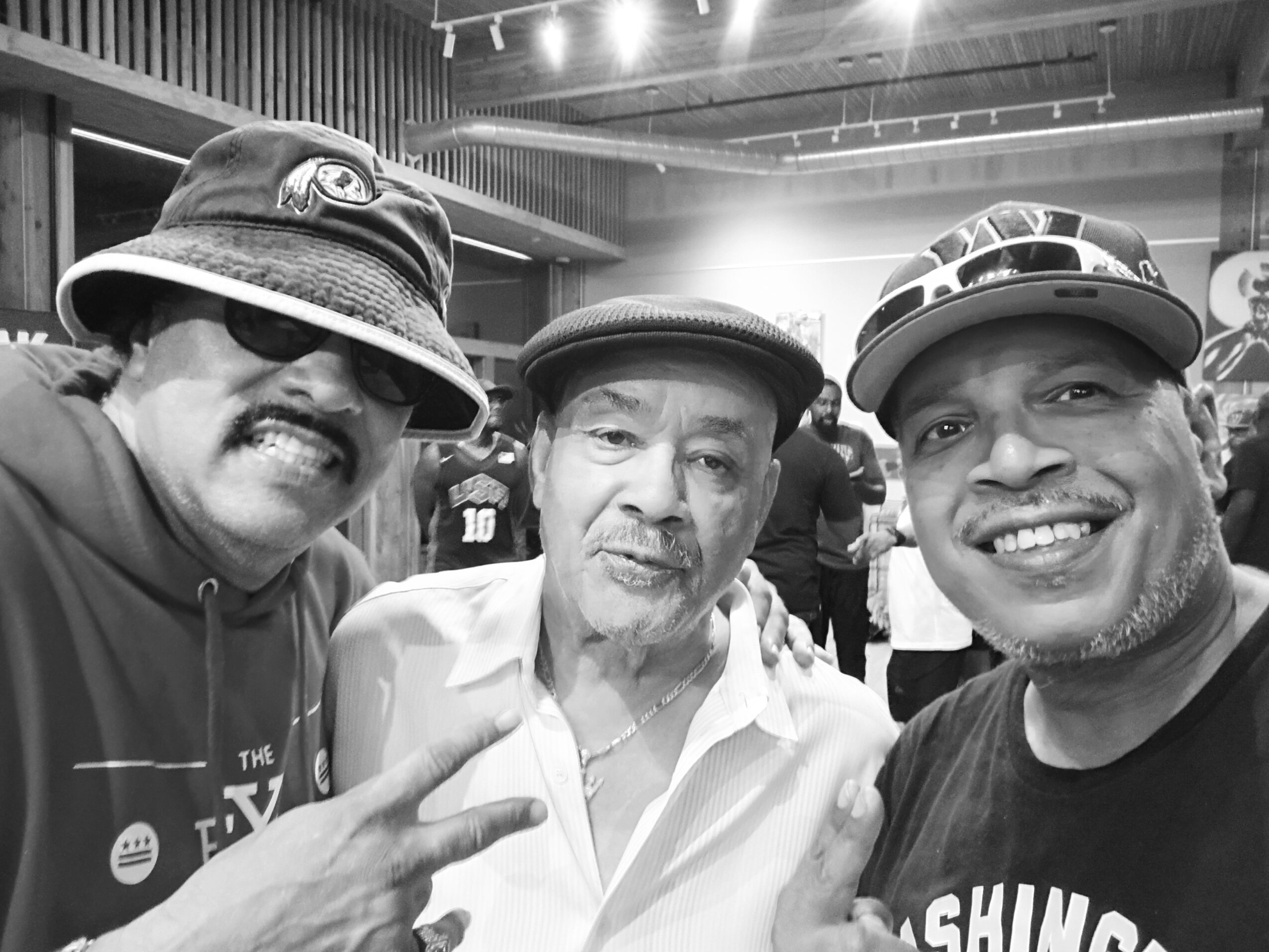 Three men smiling and posing for a photo indoors, one making a peace sign, in black and white.