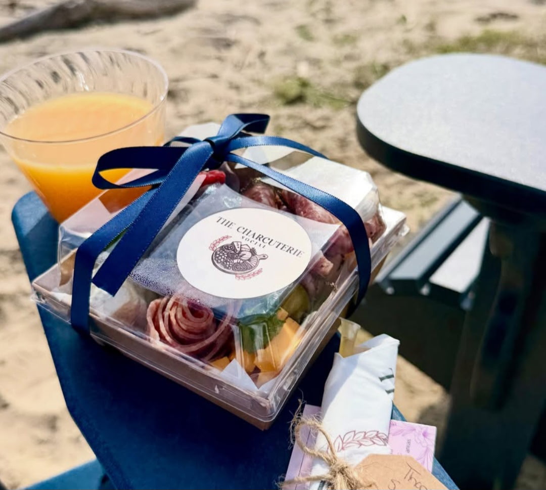 Box of assorted desserts with a blue ribbon, a glass of orange juice, and a table outdoors on sand.