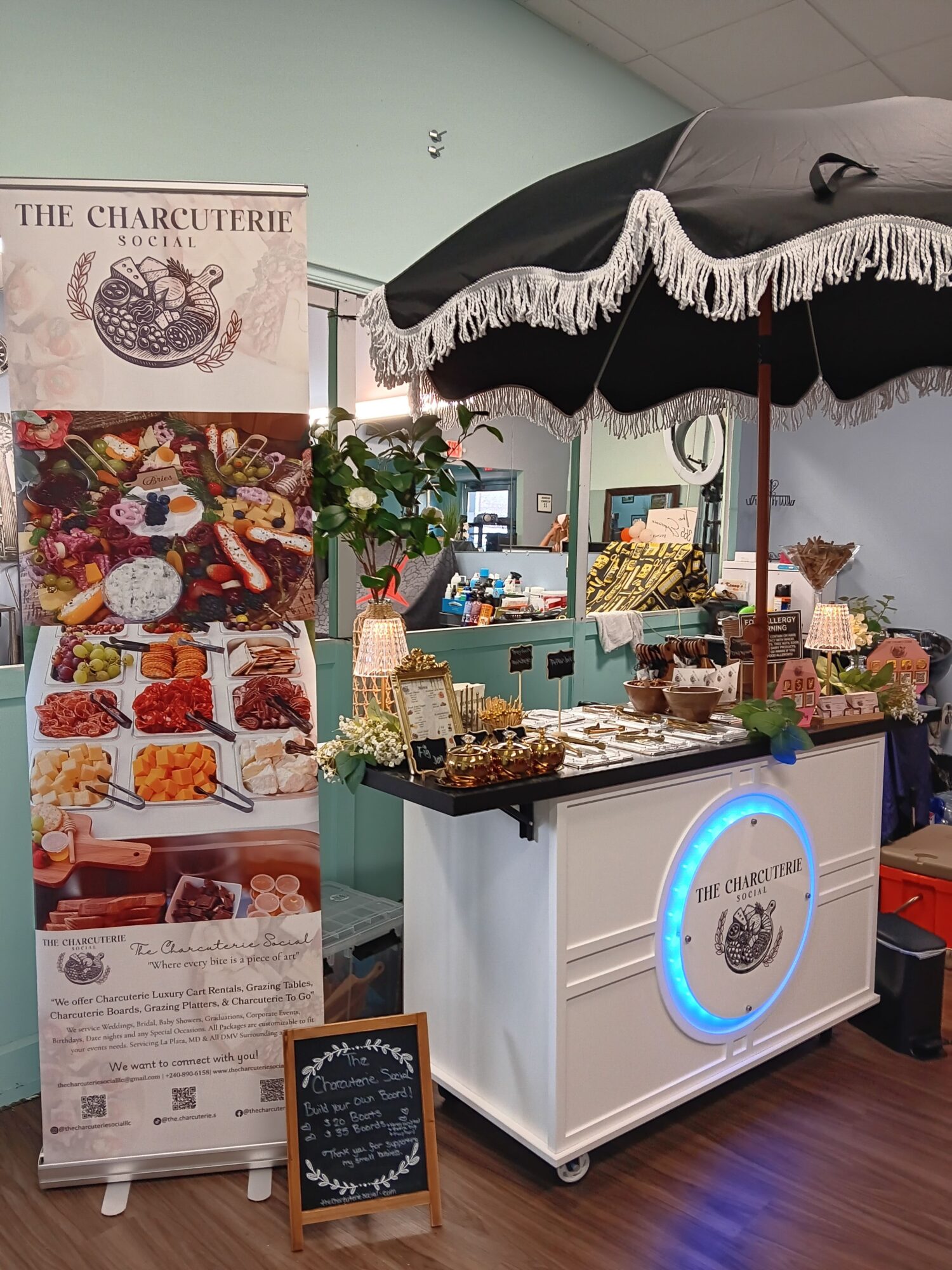 Display table with food, flowers, and a sign, under a black and white umbrella, in a room with wooden floor.