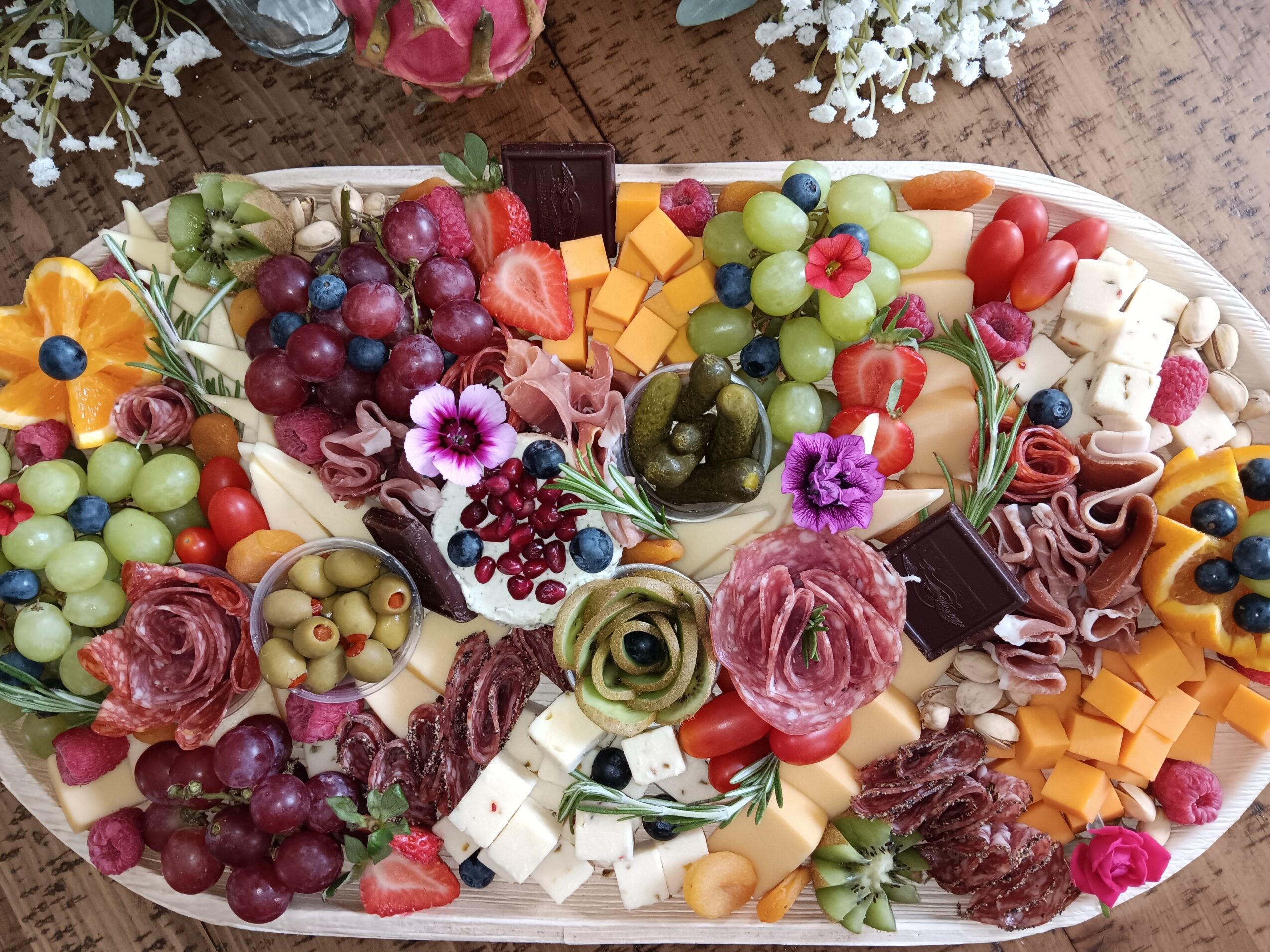 Assorted cheeses, grapes, olives, and flowers on a platter, with various colorful fruits and garnishes.