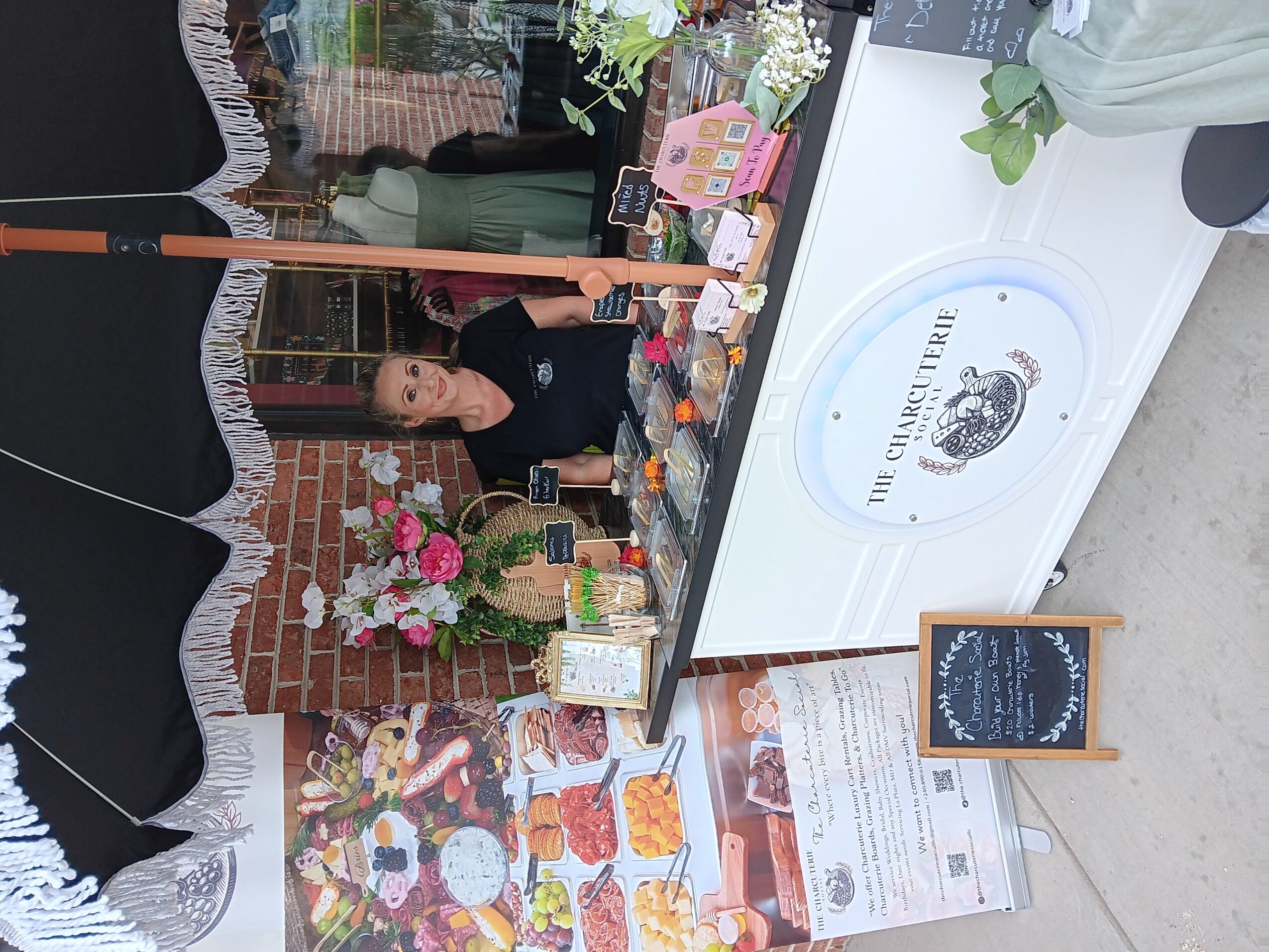 Woman sitting at a table with flowers, books, and a display, under a black umbrella, brick wall background.