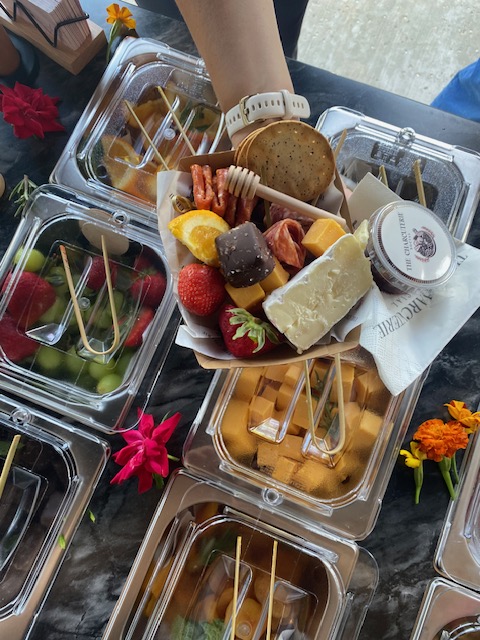 A hand holding a plate of assorted desserts and fruits, with ice cream containers and flowers on a table.