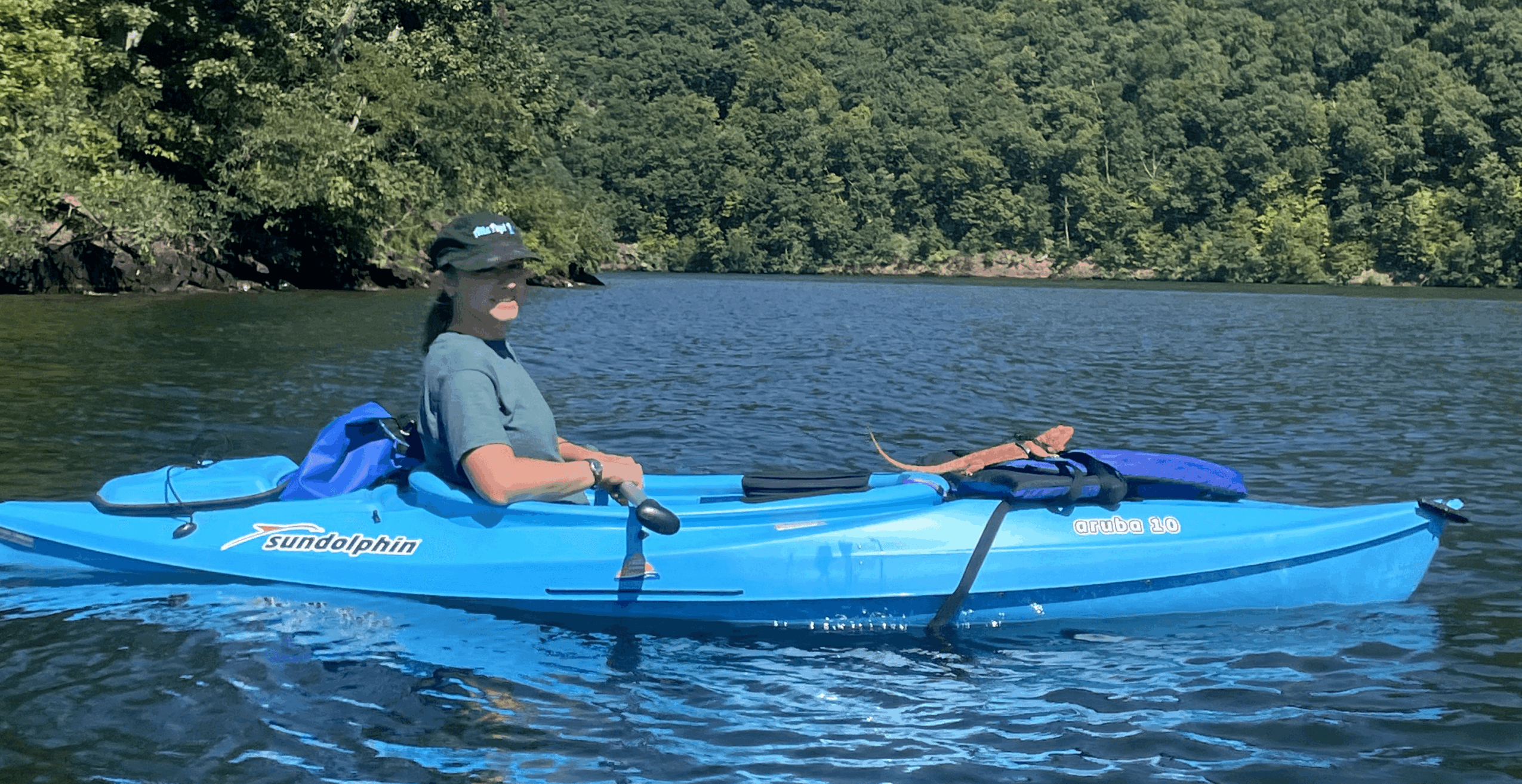 Person kayaking on a lake with trees in the background, wearing a hat and sunglasses, paddling a blue kayak.