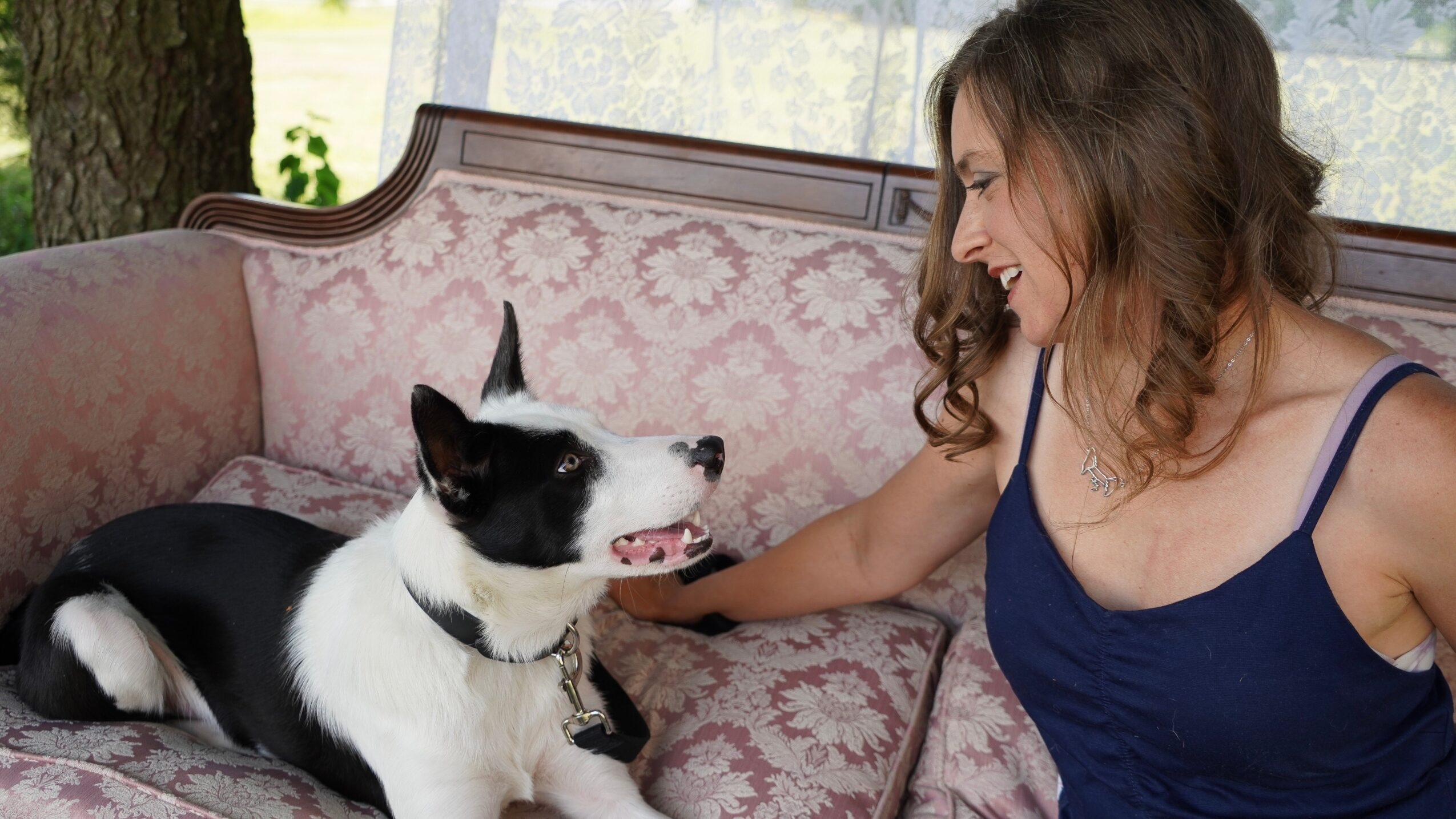 Woman with curly hair smiling at a black and white dog on a pink floral sofa indoors.