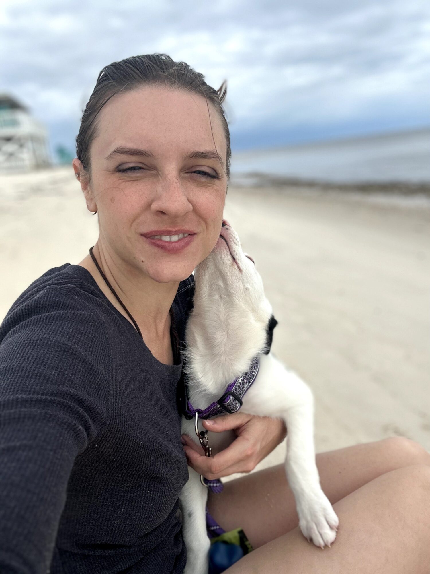 Woman taking selfie on beach with dog, cloudy sky, sandy shore, ocean in background.
