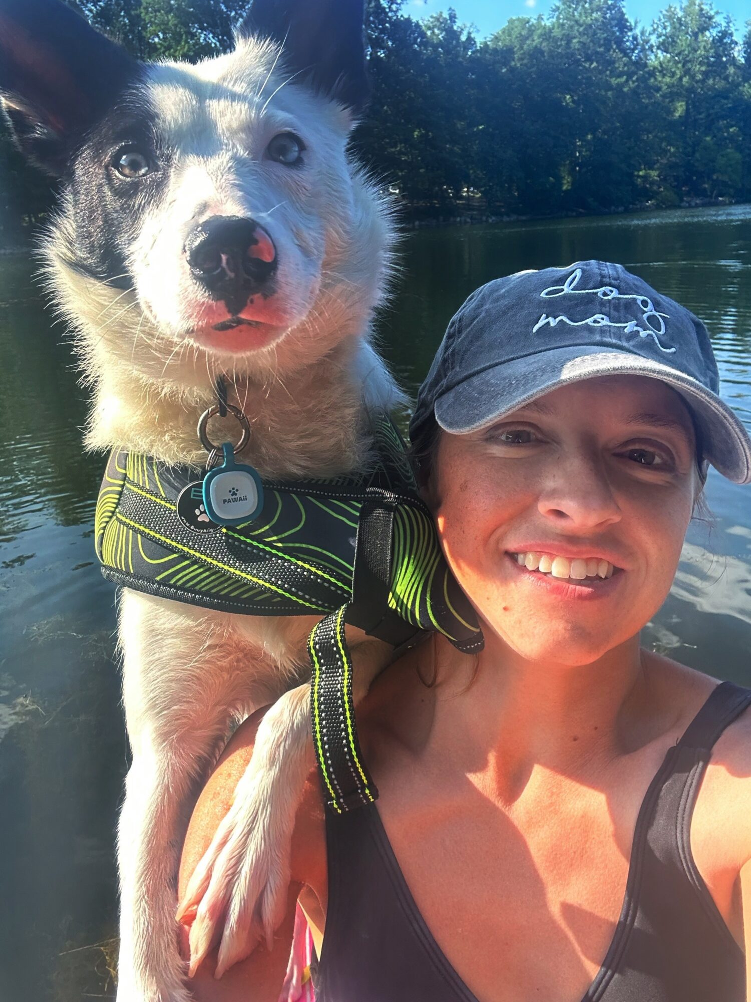 Woman smiling with a dog on her shoulder outdoors near water, wearing a cap and tank top.