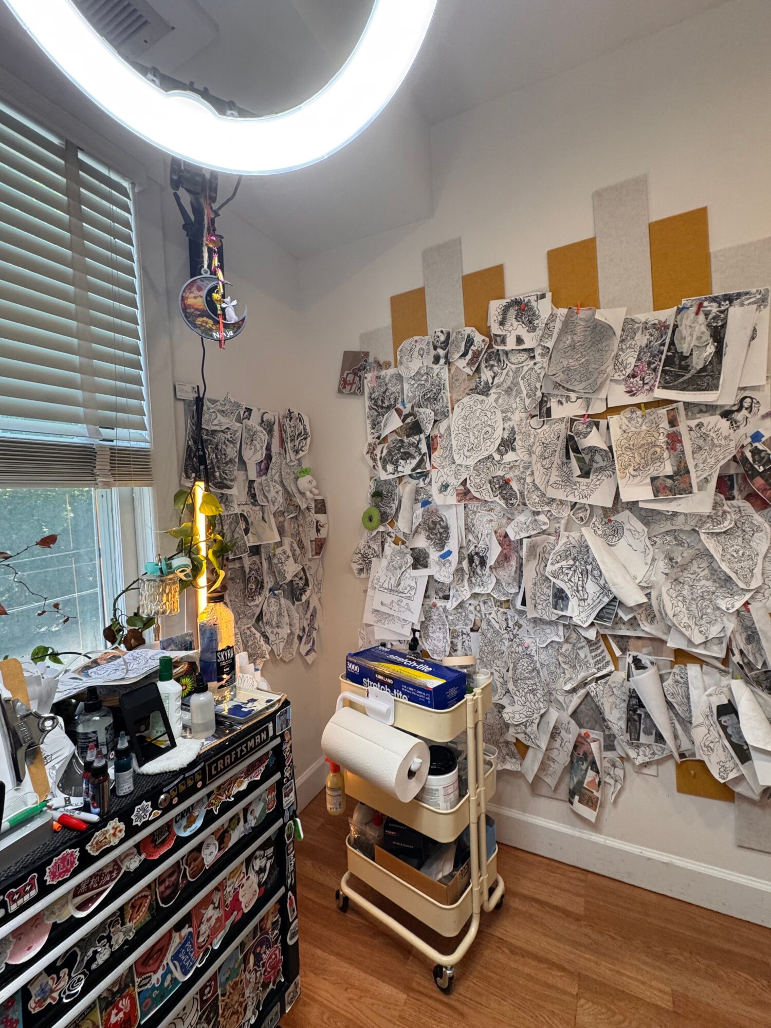 Room with a wall covered in newspaper clippings, a window with blinds, and various objects on a table and shelves.