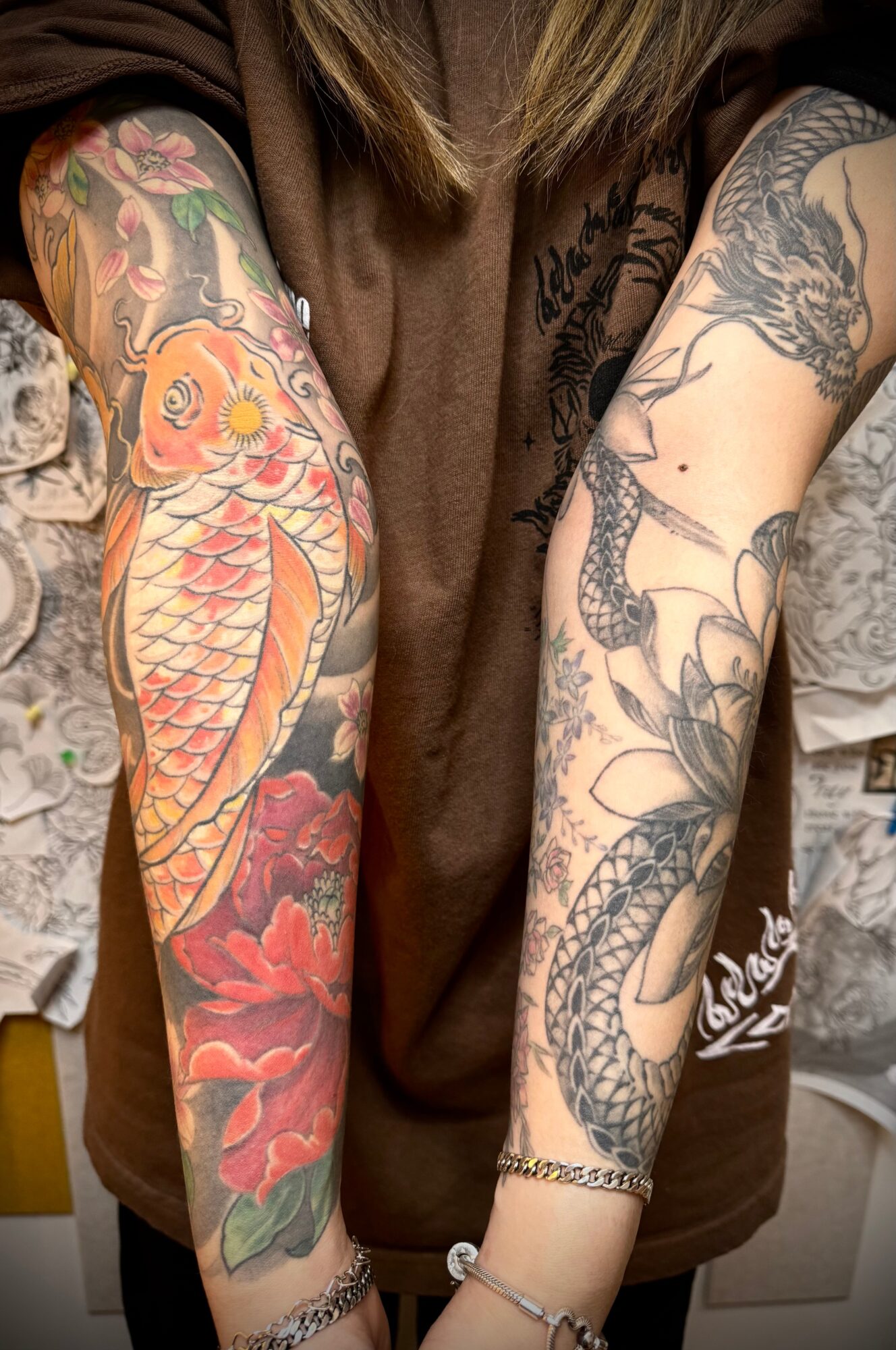 Two tattooed arms with colorful and black ink designs, including a koi fish and floral patterns, held close together.