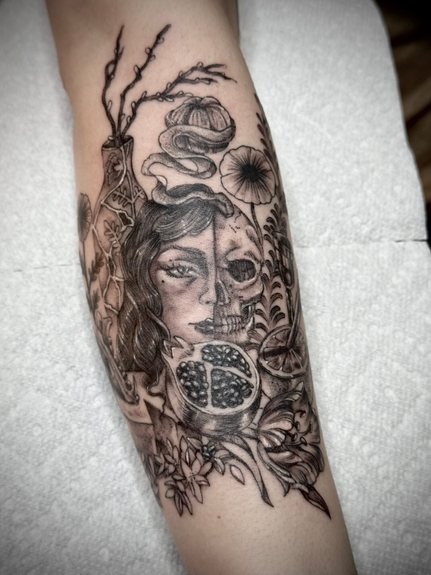 Black and gray tattoo of a woman's face, a tree, a snake, a pomegranate, and various plants on arm.