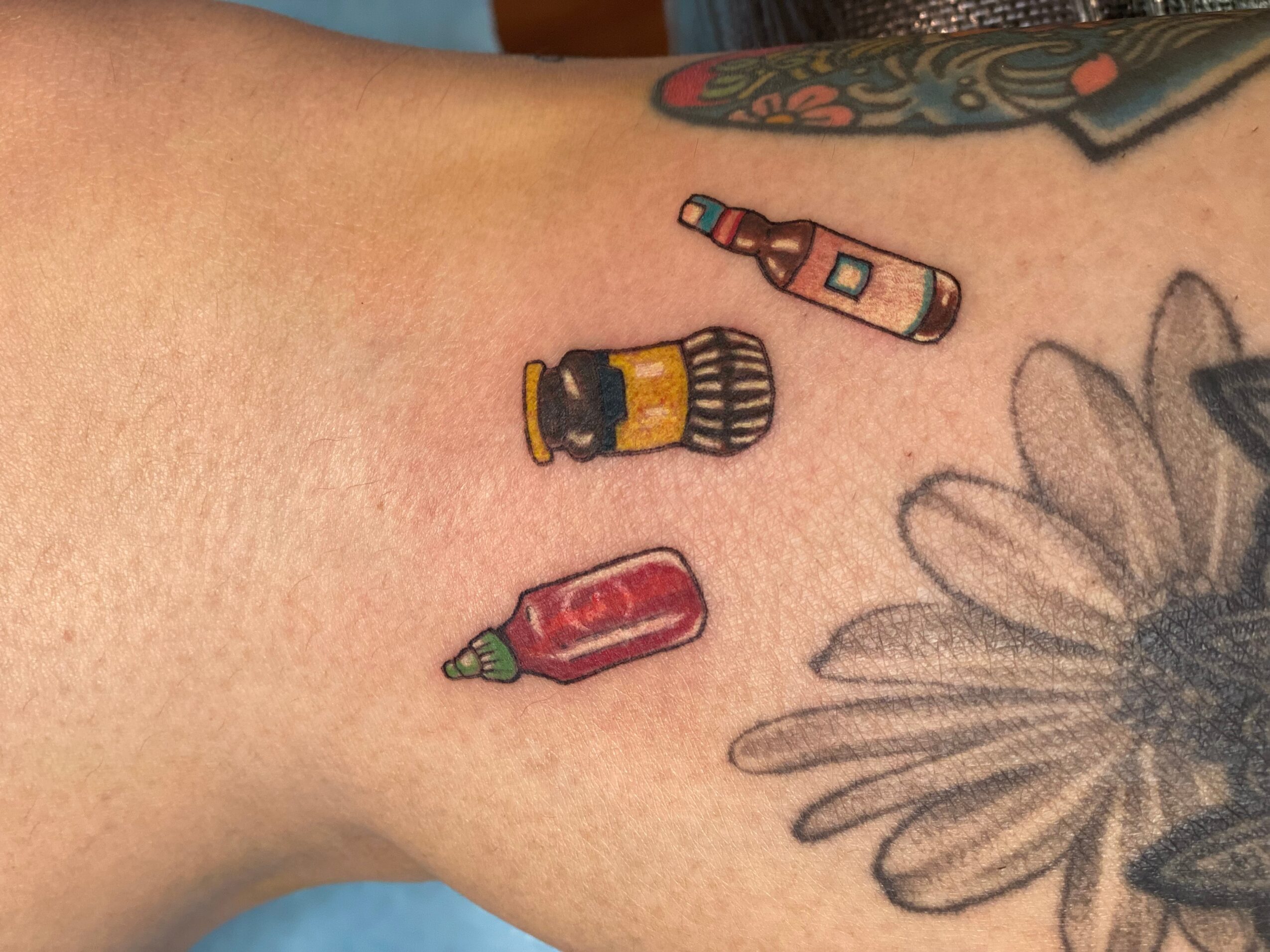 Tattoo of a salt shaker, ketchup bottle, and hot sauce bottle on skin, with a flower tattoo partially visible nearby.