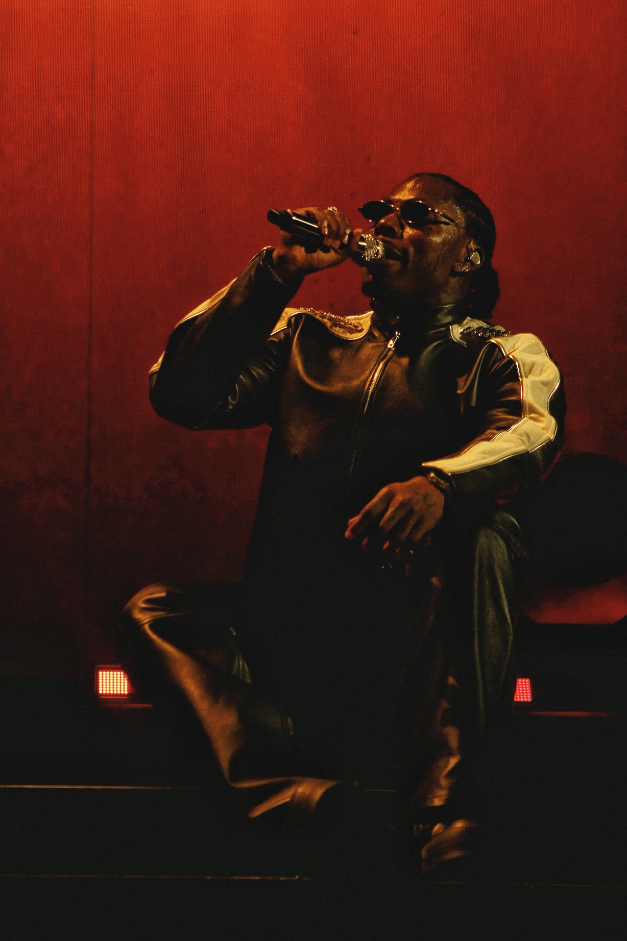 Person singing into microphone on stage with red background, wearing sunglasses and dark clothing.