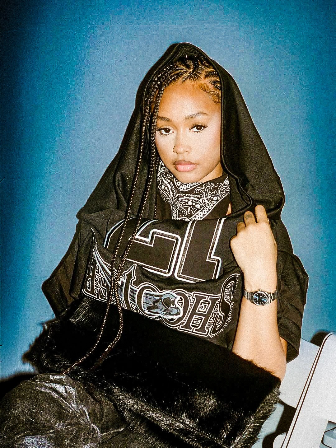 Young woman with braided hair wearing a black hoodie and patterned bandana, holding a black bag, against a blue background.
