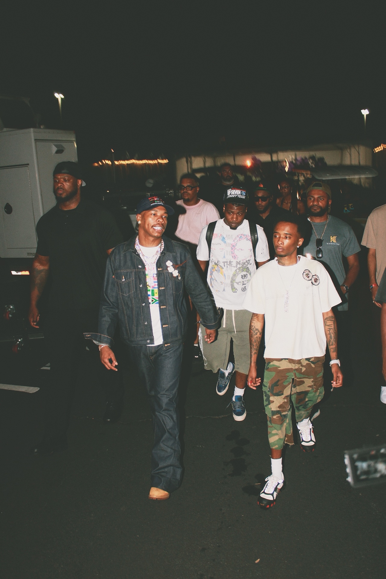 Group of people walking outdoors at night, some wearing casual clothing, with a dark background and lights in the distance.