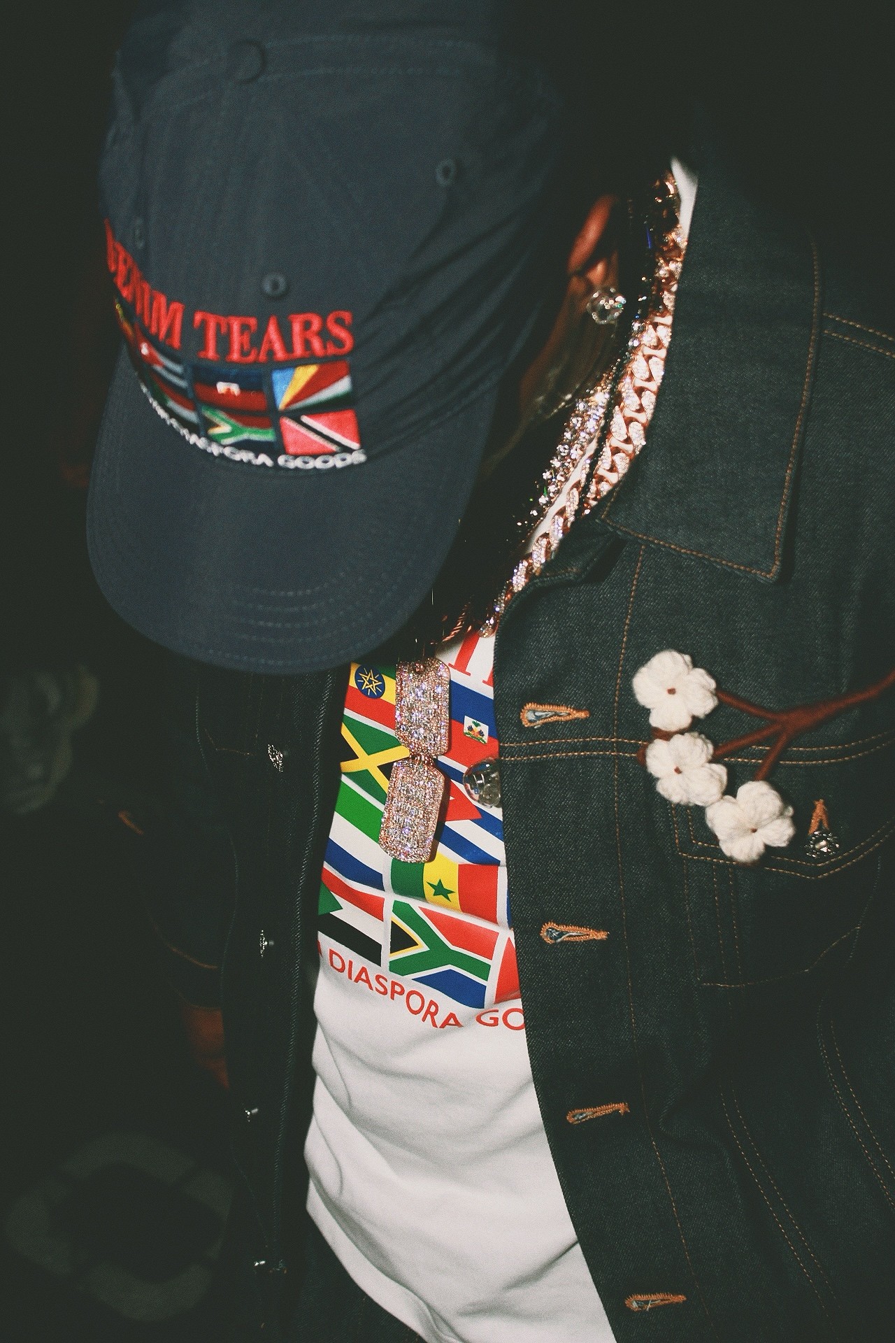 Person wearing a cap, a jacket, and a shirt with various flags, with a beaded necklace, in a dark setting.