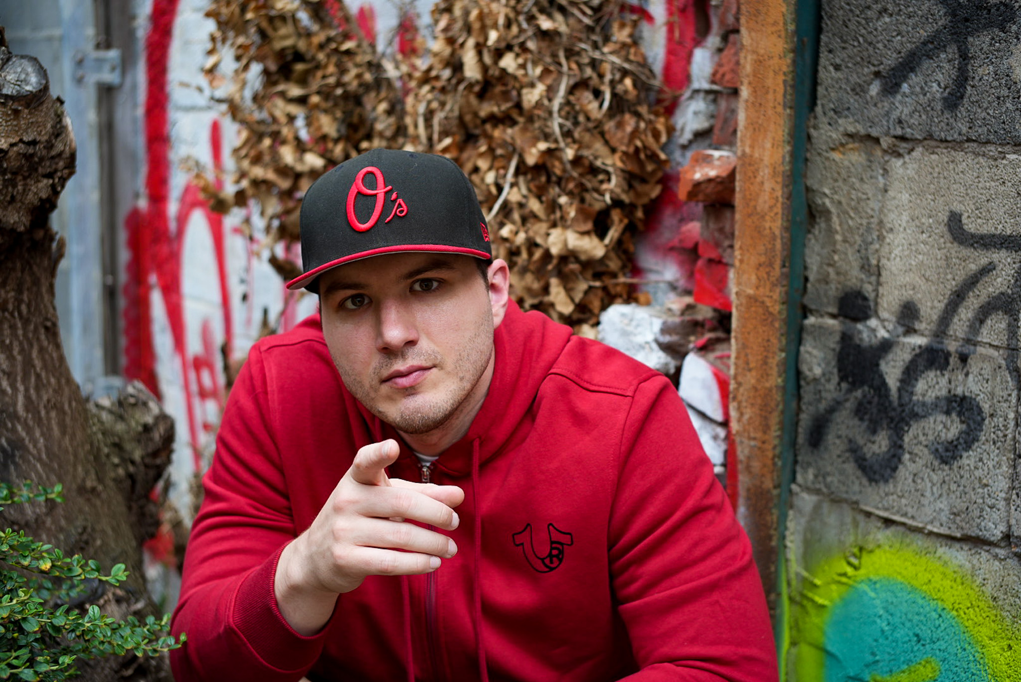 Young man wearing a black cap and red hoodie, pointing at the camera, outdoors with graffiti and dried leaves behind him.