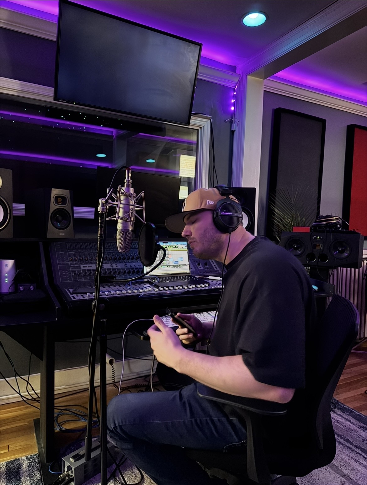 Man wearing headphones in a recording studio, holding a phone, with audio equipment and a large monitor behind him.