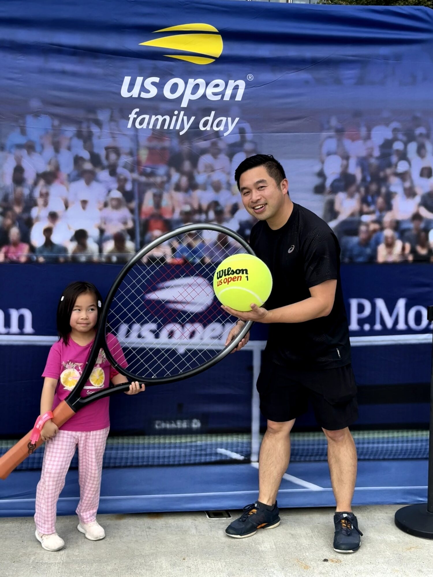 A young girl and a man holding a tennis racket and a yellow tennis ball at US Open Family Day, with a crowd background.