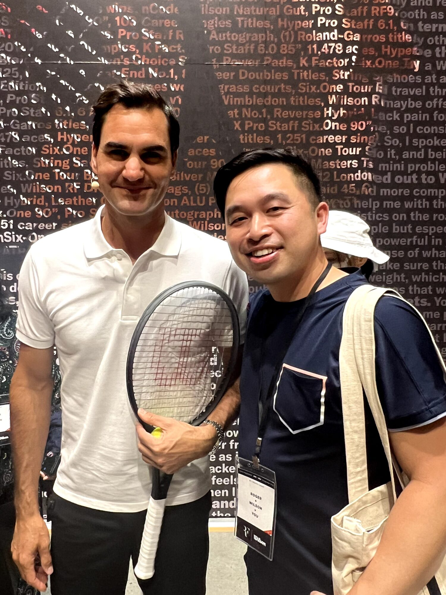 Two men smiling, one holding a tennis racket, standing in front of a wall with text, one wearing a white shirt, the other a dark shirt with a backpack.