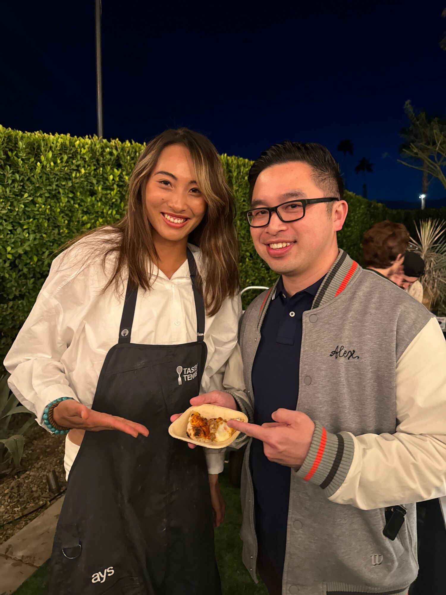 Two people standing outdoors at night, one holding a taco, smiling, with greenery in the background.