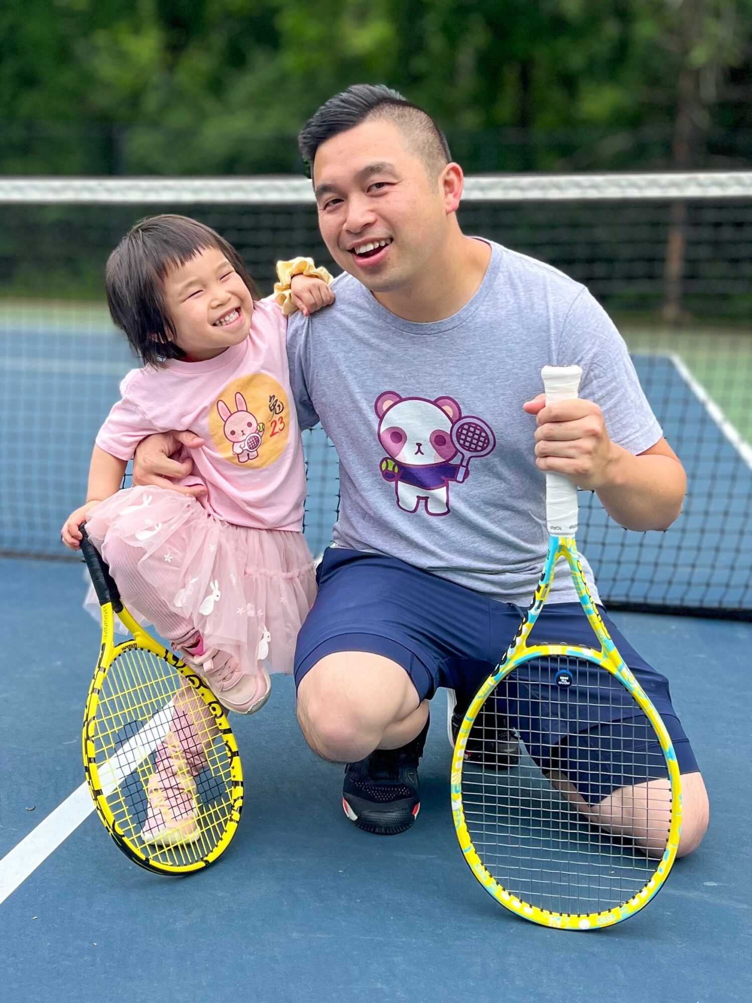 Man and young girl smiling on tennis court, holding tennis rackets, with tennis ball and drink, outdoors, trees in background.