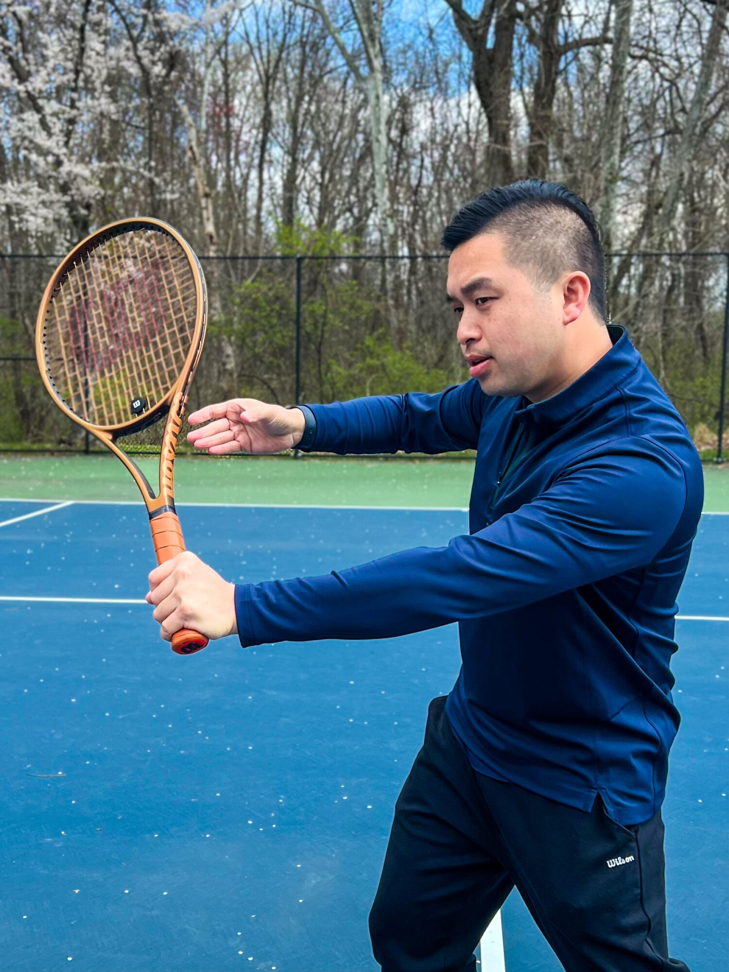 Man in blue jacket holding tennis racket on outdoor court, trees in background.