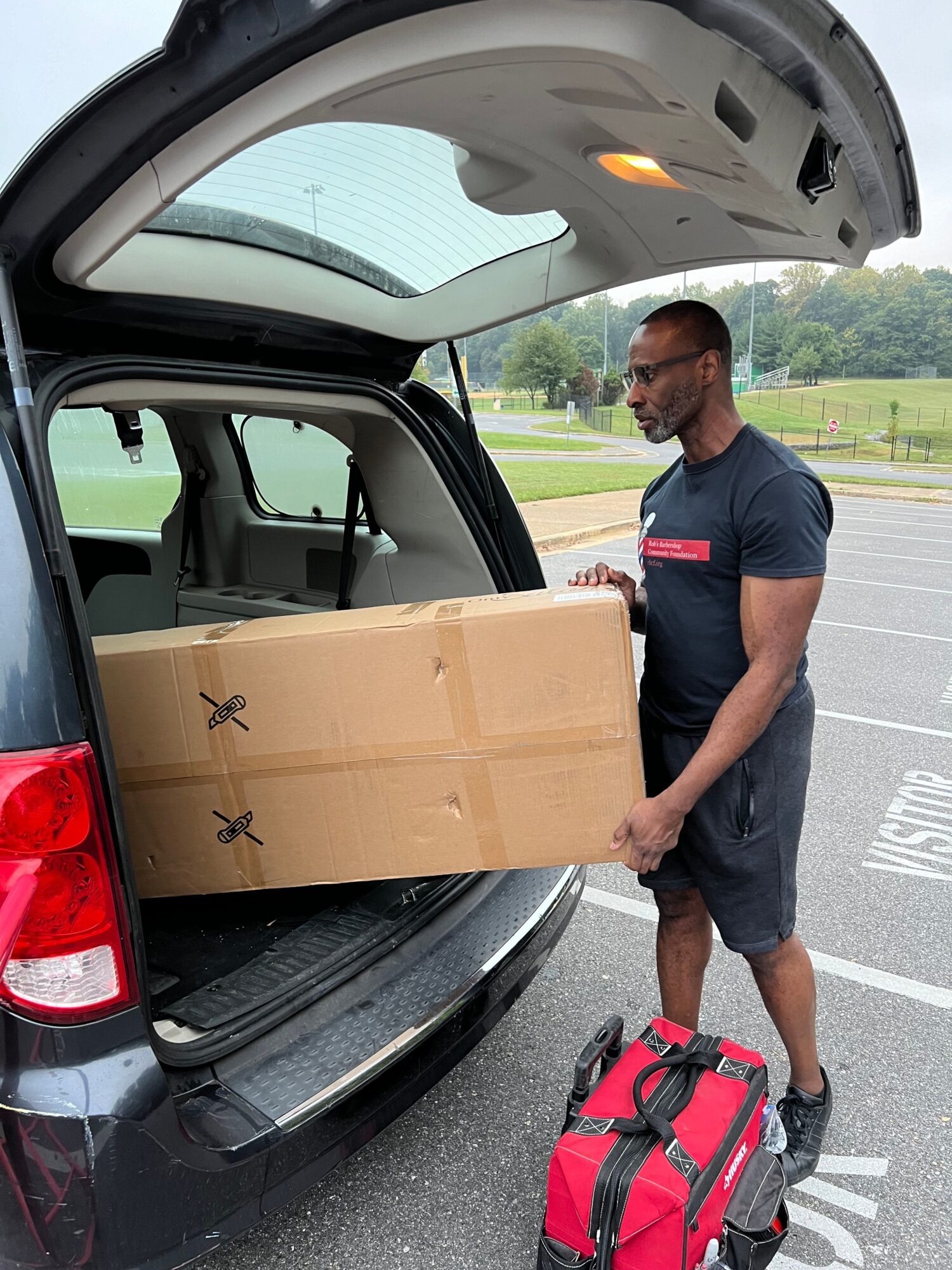 Person standing next to a car with an open trunk, holding a large cardboard box, in a parking lot.
