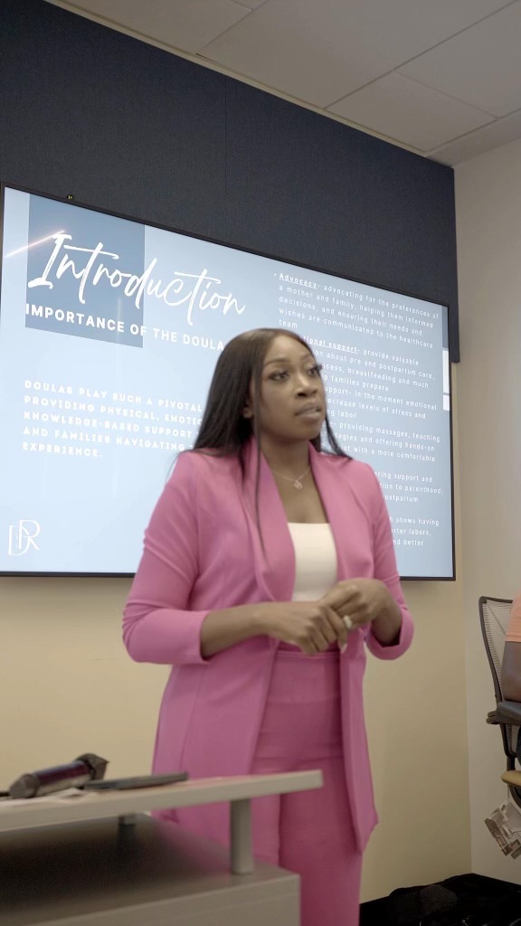 Woman in pink blazer and pants standing in front of a presentation screen, holding her hands together, in a classroom or conference room.