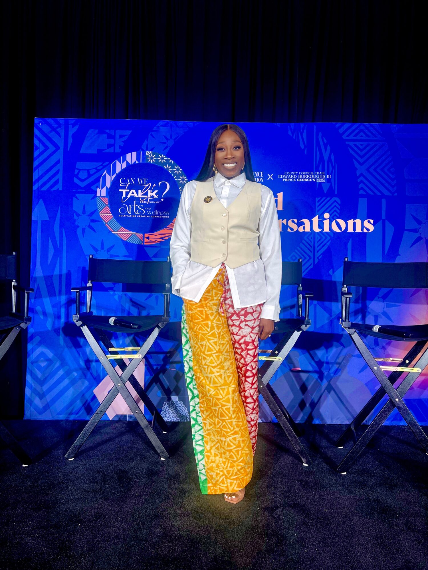 Woman standing on stage with blue background, smiling, wearing colorful pants and a white jacket, chairs behind her.