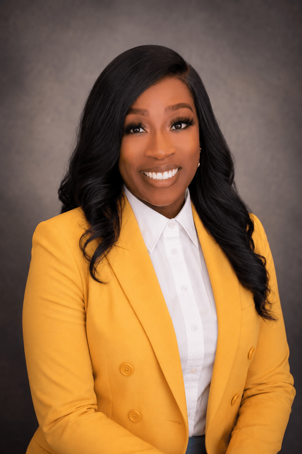 Smiling woman with long black hair in a yellow blazer and white shirt, against a gray background.