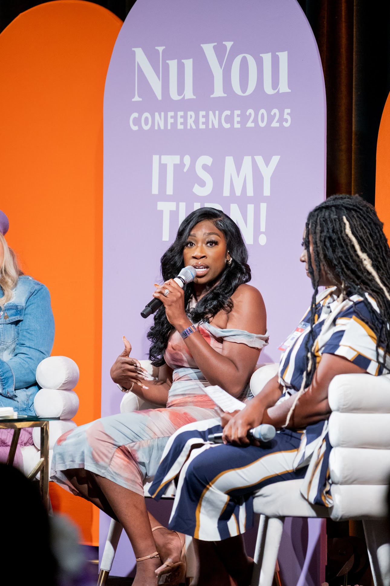 Three women seated on stage, one speaking into a microphone, with a purple banner behind them reading NuYou Conference 2025.