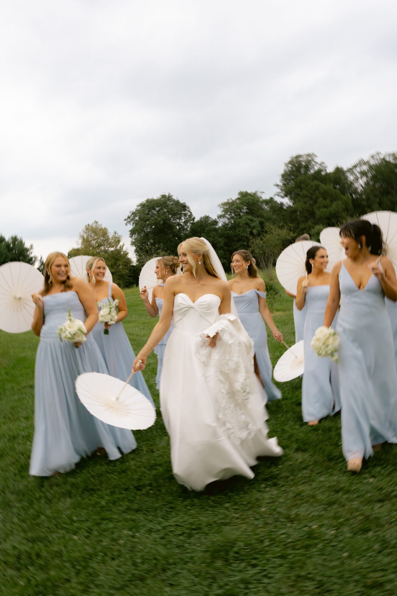 Bride in white dress walking with bridesmaids in light blue dresses holding umbrellas and flowers outdoors on grass under cloudy sky.