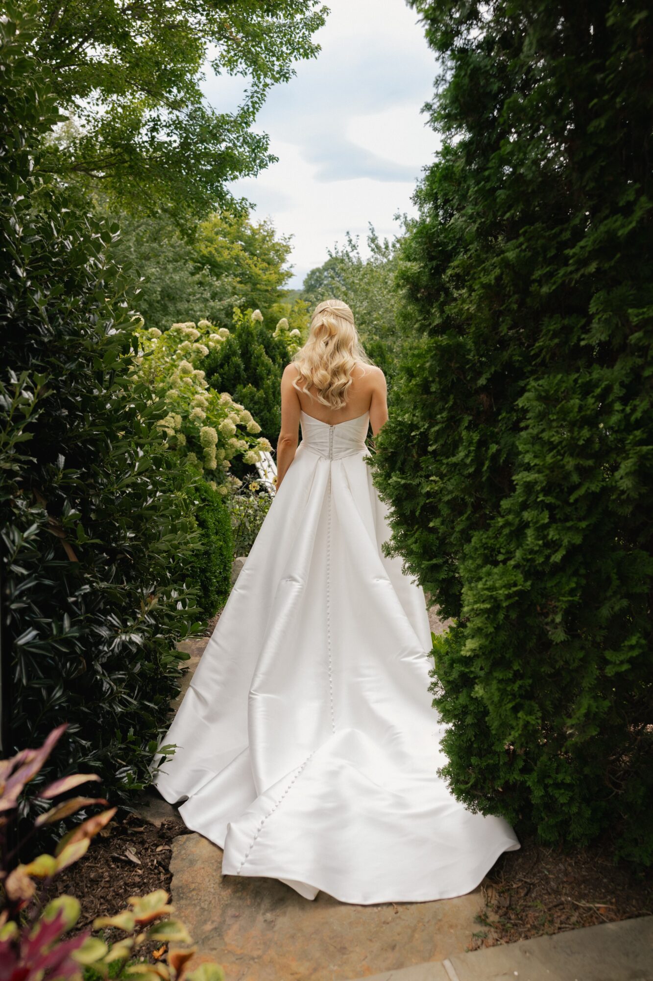 Woman in a white wedding dress walking away through greenery, back view, outdoors, daytime.