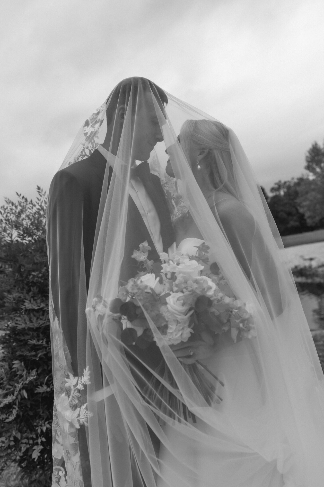 Two people with wedding veils face each other, holding a bouquet, outdoors with trees and sky in background.