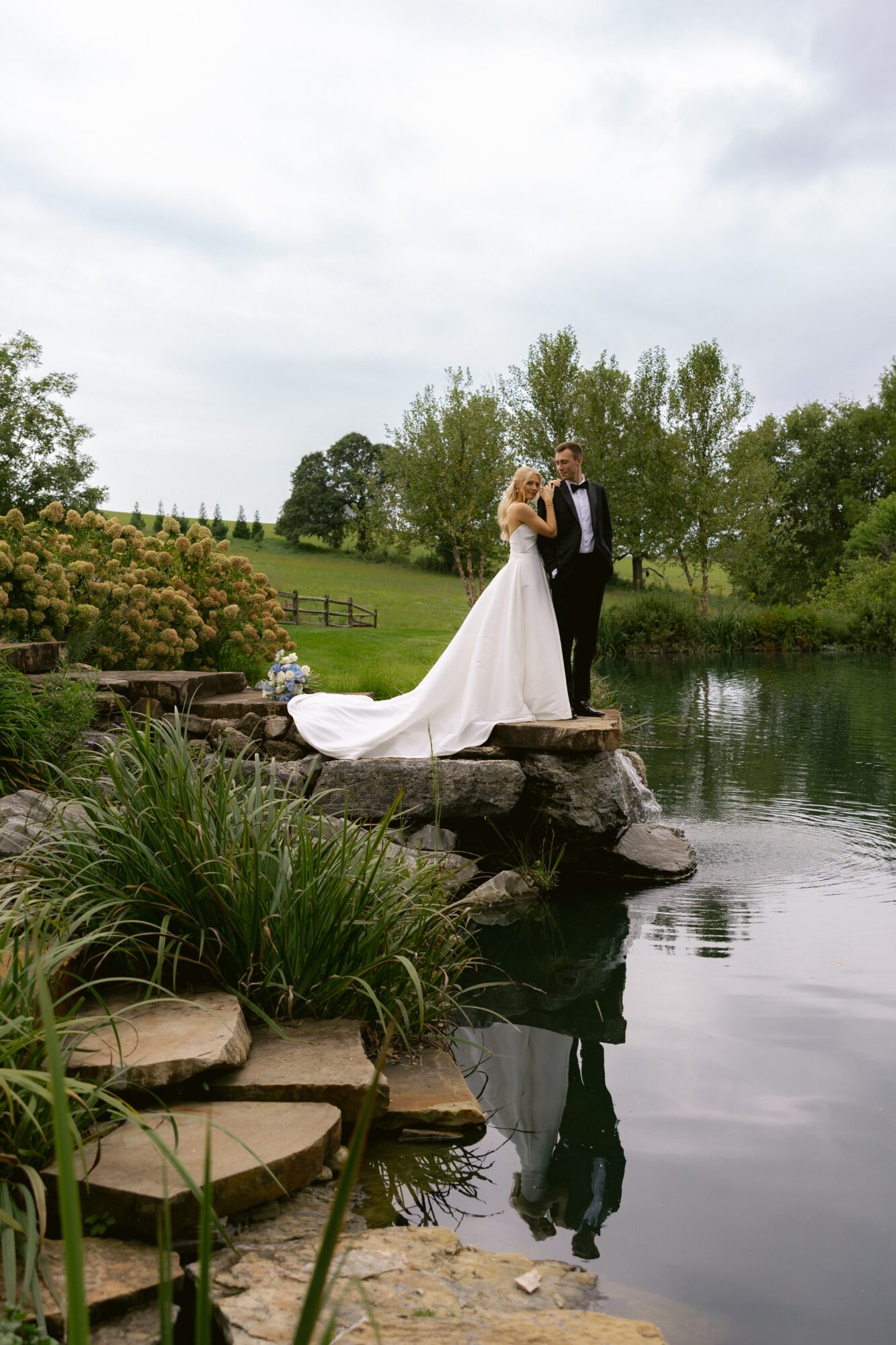 Bride and groom standing on rocks by a pond, surrounded by greenery and trees, with their reflection in the water.