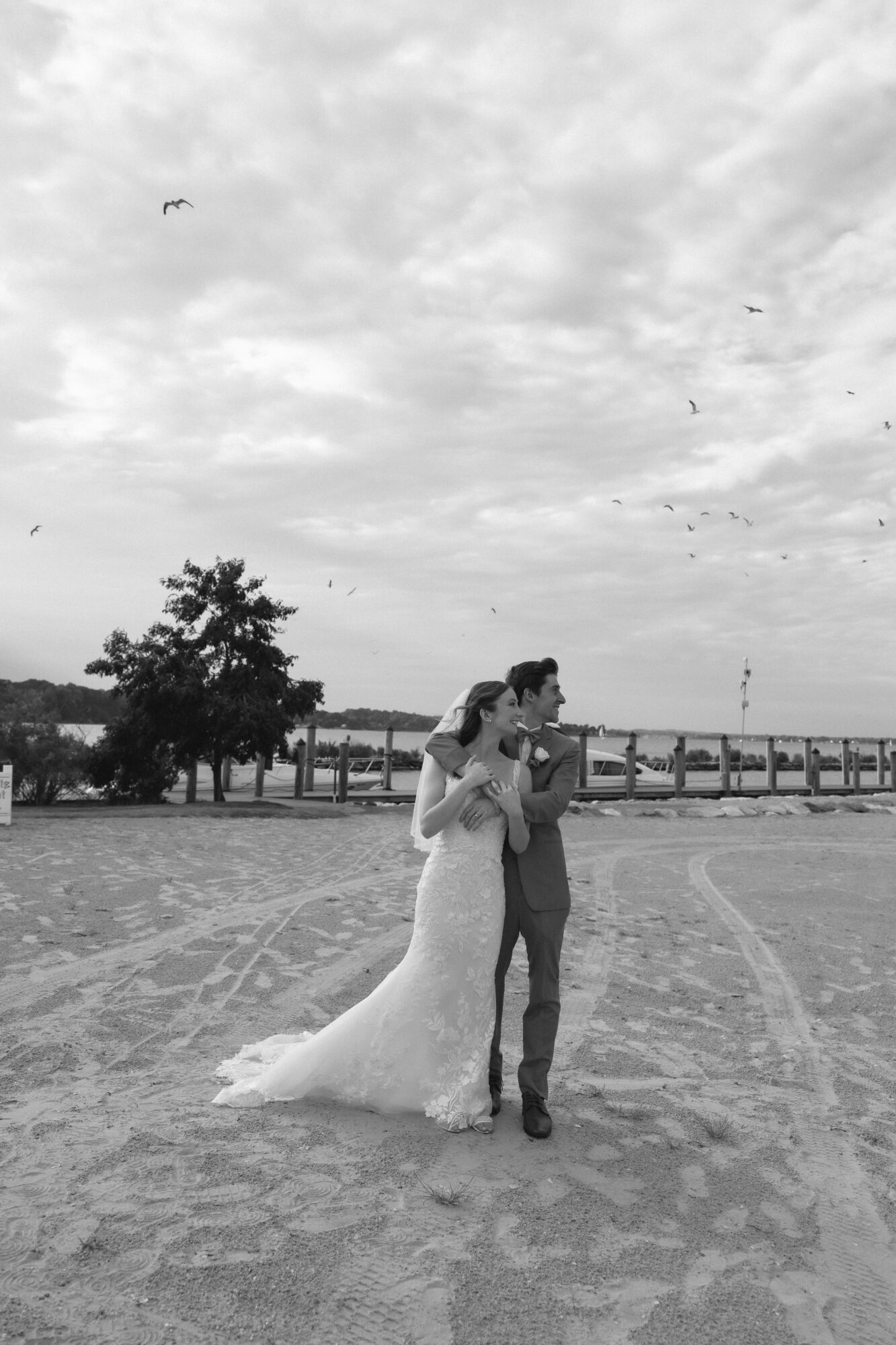 A bride and groom stand together outdoors on a cloudy day, with a body of water and trees in the background.