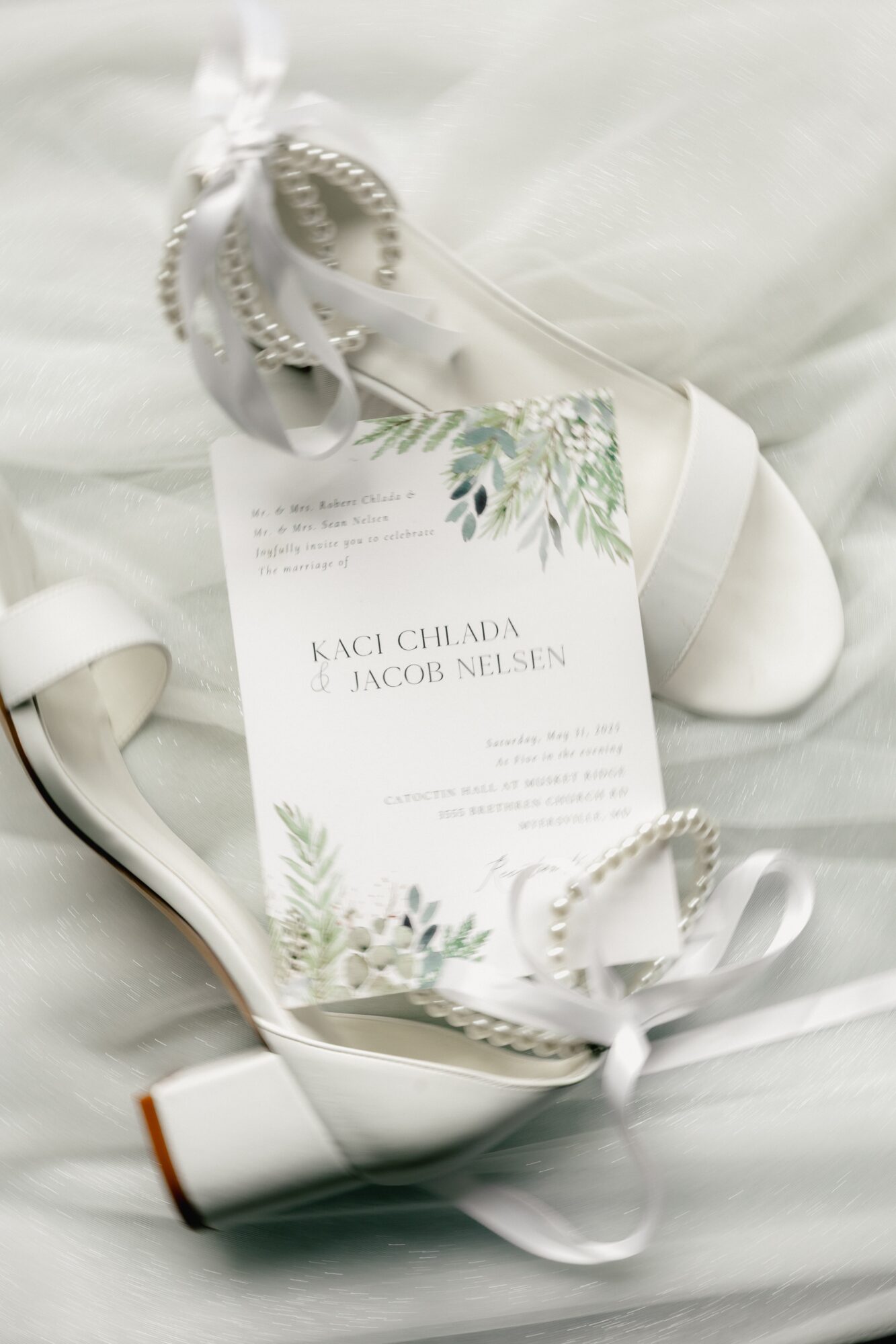 Wedding invitation card with floral design, placed inside a white shoe with ribbons, on a soft fabric background.