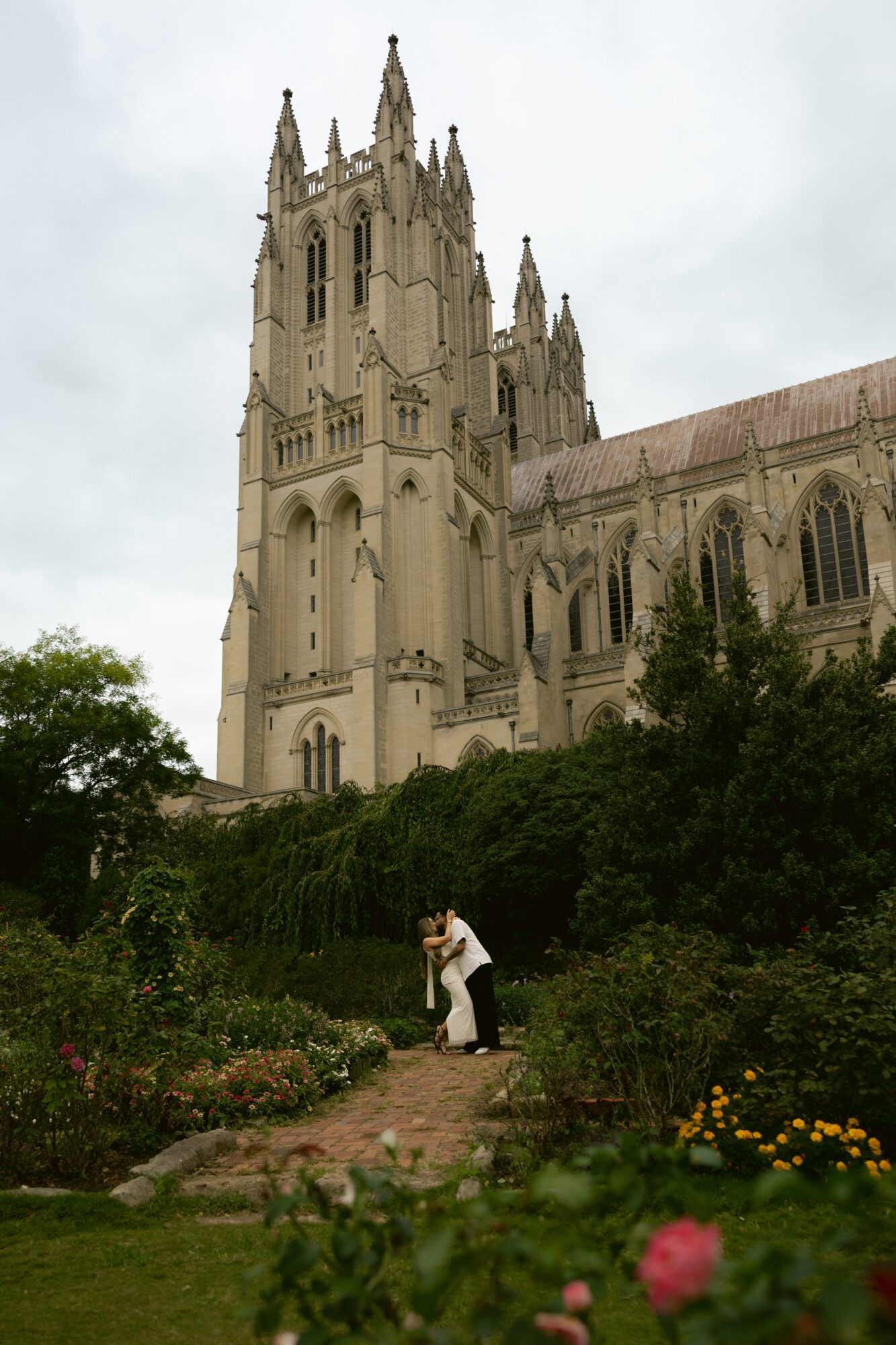 A person in a garden with a large cathedral in the background, cloudy sky, greenery, and flowers.