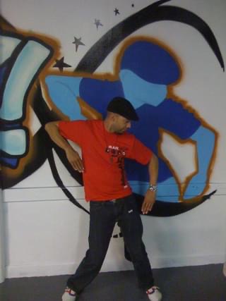 Young person in red shirt and black pants standing in front of a colorful graffiti wall, looking down.